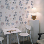 A small dining area with a white table and chairs against a wall covered in dreamy orchid wallpaper. The wallpaper showcases a delicate pattern of blue orchids, soft pink roses, and small sprigs of baby's breath on a light background, creating a calming and inviting atmosphere. A table lamp on a white pedestal adds a warm glow to the space.