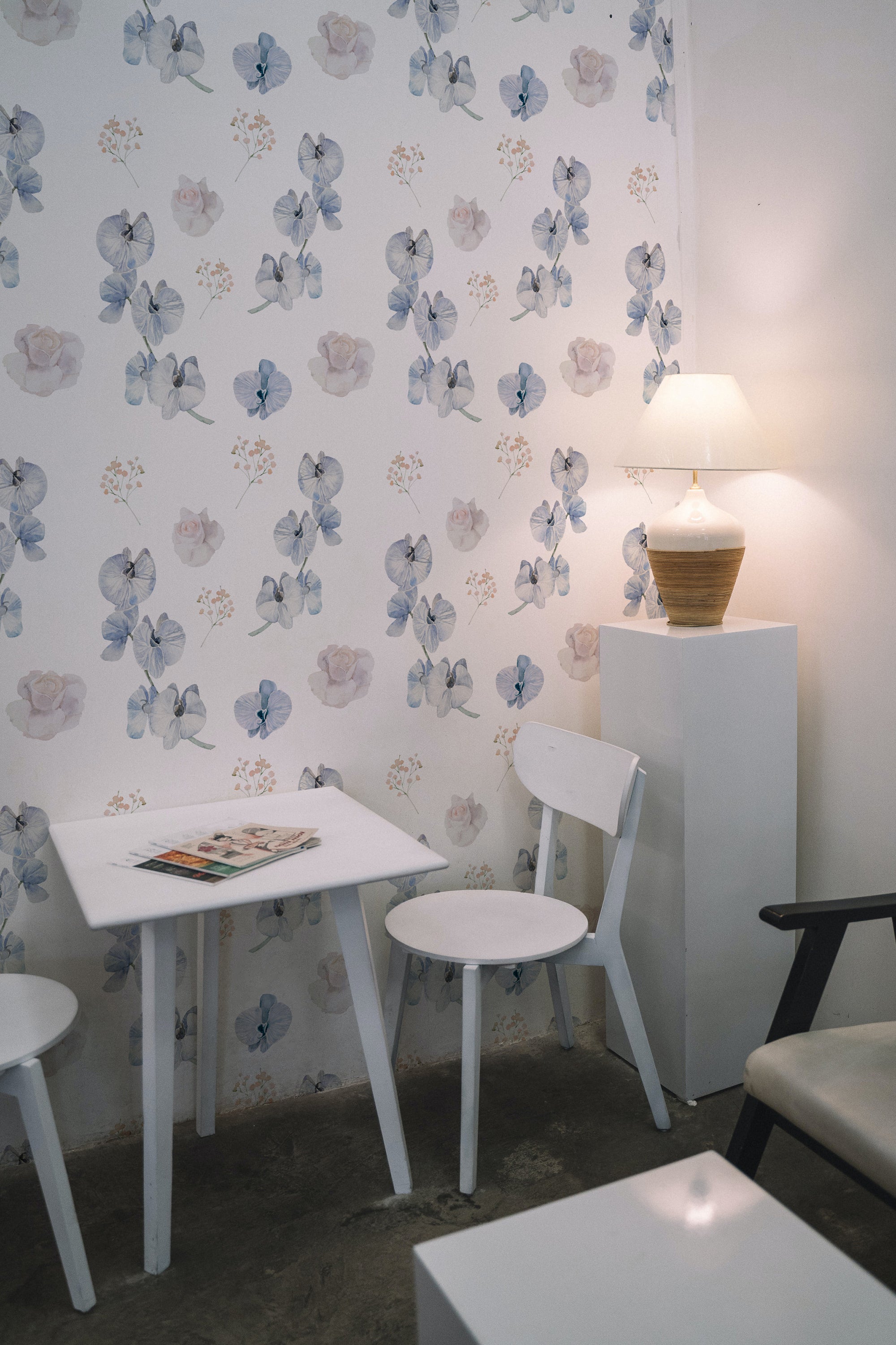 A small dining area with a white table and chairs against a wall covered in dreamy orchid wallpaper. The wallpaper showcases a delicate pattern of blue orchids, soft pink roses, and small sprigs of baby's breath on a light background, creating a calming and inviting atmosphere. A table lamp on a white pedestal adds a warm glow to the space.