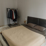 A modern bedroom featuring walls decorated with Fabric 10B Wallpaper, showcasing vertical stripes in soothing beige and soft blue tones, complemented by a minimalistic bed and a clothing rack.