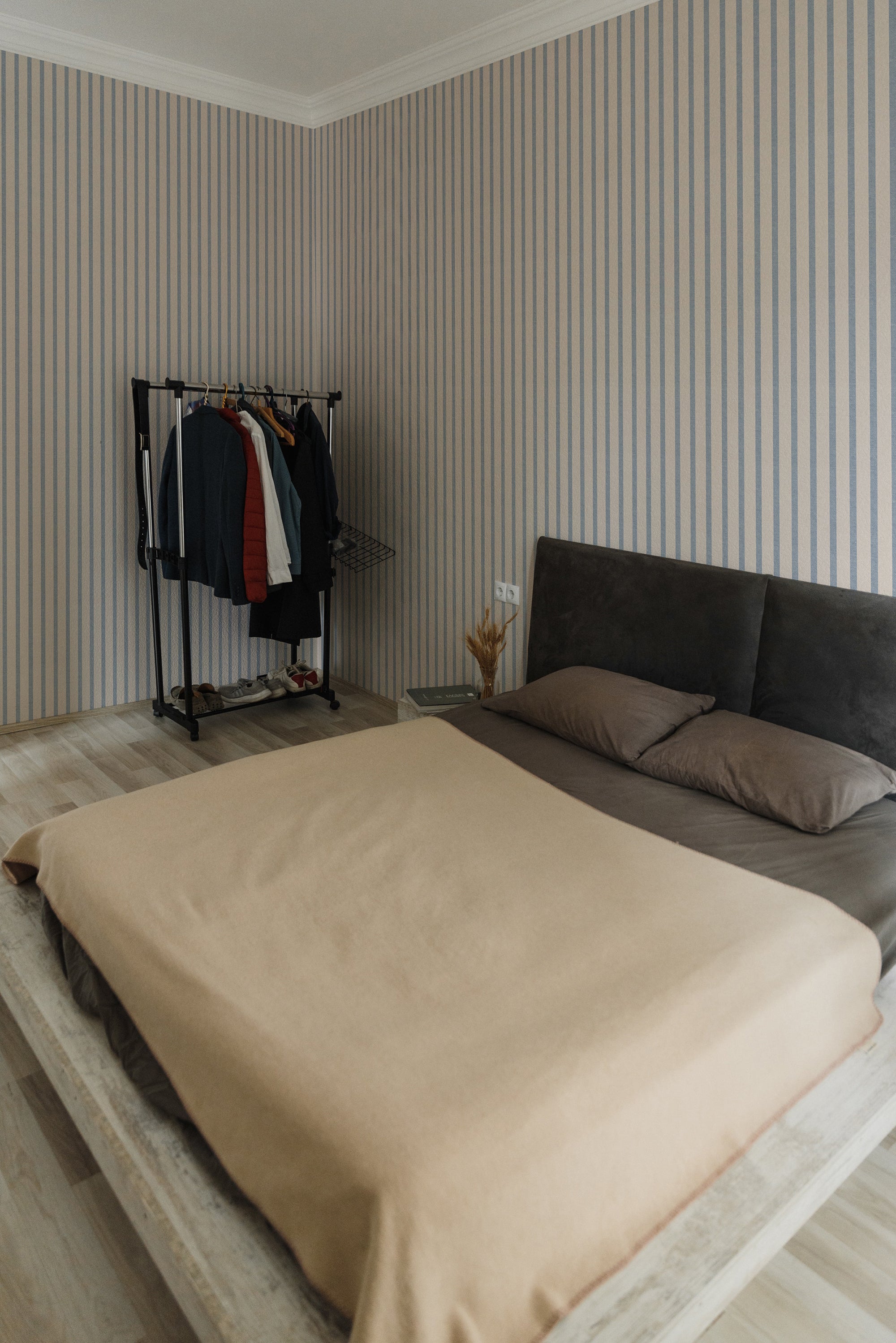 A modern bedroom featuring walls decorated with Fabric 10B Wallpaper, showcasing vertical stripes in soothing beige and soft blue tones, complemented by a minimalistic bed and a clothing rack.