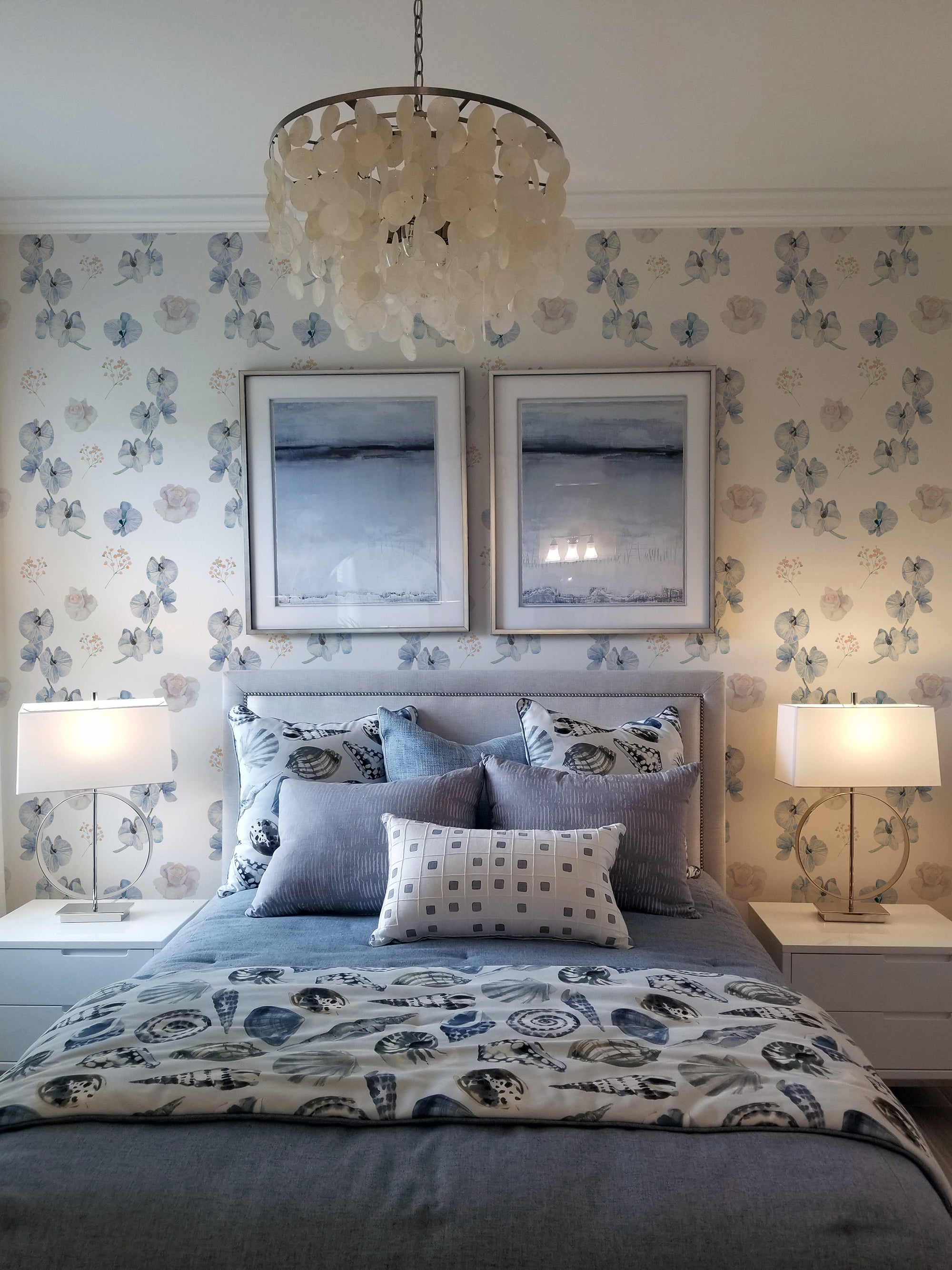 A cozy bedroom with a bed and nightstands against a wall covered in dreamy orchid wallpaper. The wallpaper features a delicate design of blue orchids, soft pink roses, and small sprigs of baby's breath on a light background, adding a touch of elegance and serenity to the room. The bed is adorned with blue and white bedding and framed artwork hangs above the headboard.