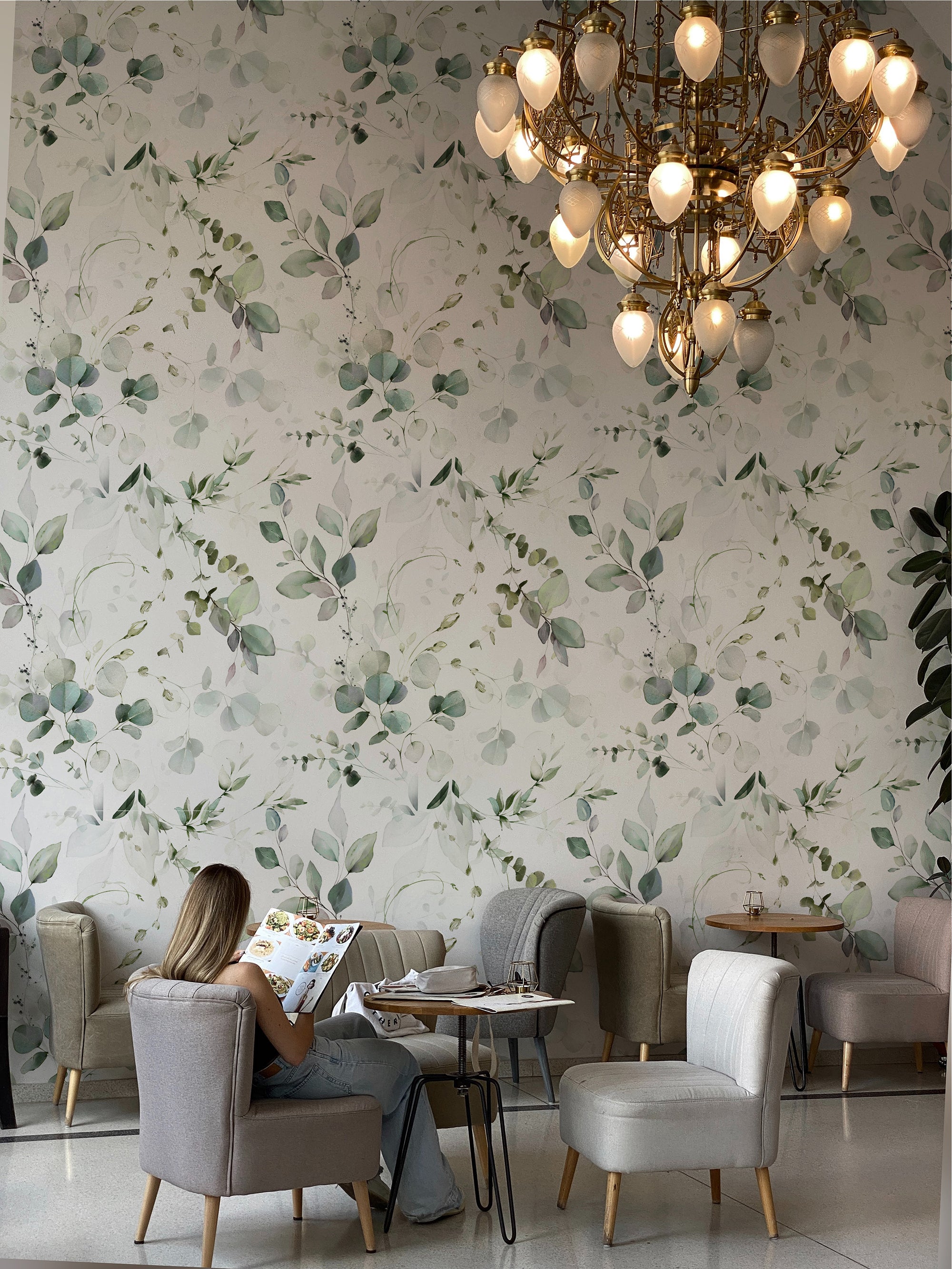 An elegant cafe interior showcasing the Eucalyptus Wallpaper with lush green and soft grey eucalyptus leaf patterns, a woman seated reading a menu, under the warm glow of a sophisticated golden chandelier.