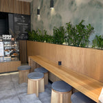 Interior of a modern café with a Shimmering Marble Wallpaper feature wall displaying a beautiful marble effect in shades of blue and green with gold veins. The setting includes a wooden bench with grey padded stools and green plants enhancing the natural ambiance