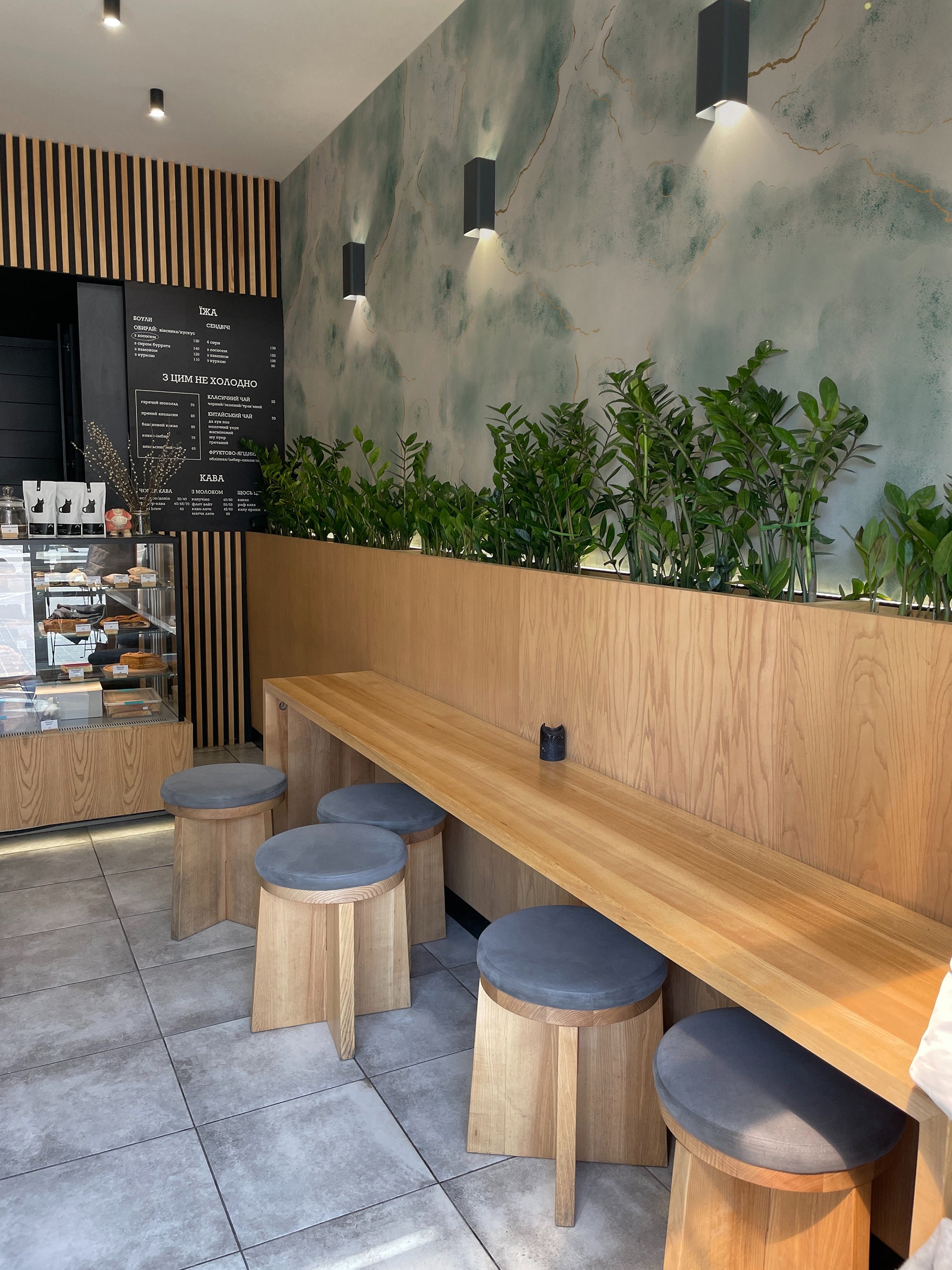 Interior of a modern café with a Shimmering Marble Wallpaper feature wall displaying a beautiful marble effect in shades of blue and green with gold veins. The setting includes a wooden bench with grey padded stools and green plants enhancing the natural ambiance