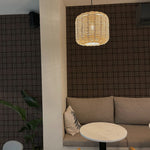 A cozy corner of a modern cafe, illuminated by a woven pendant lamp, showcasing walls covered in Dark Plaid Wallpaper - Black with a subtle black and white check pattern. The area features a comfortable seating nook with grey cushions, a round table, and a natural wood chair, creating an inviting space for patrons.