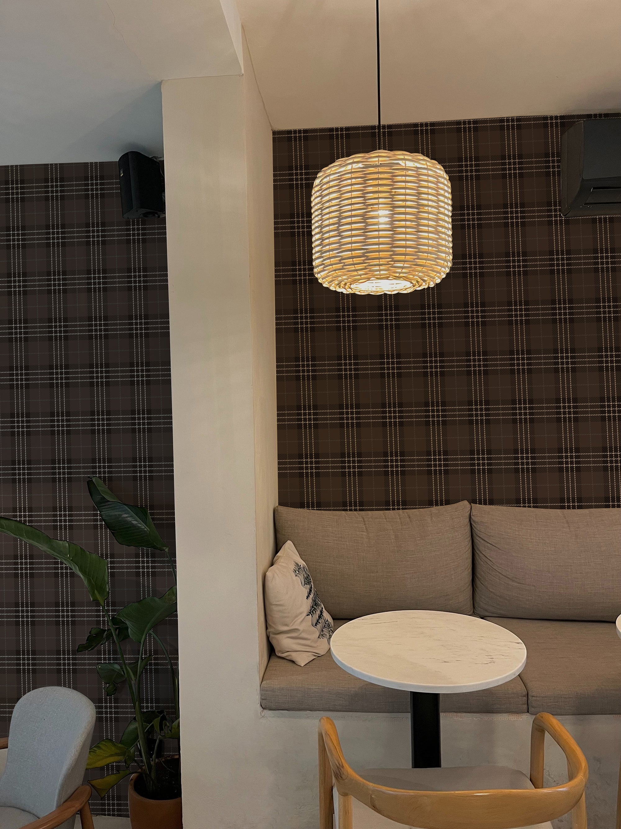 A cozy corner of a modern cafe, illuminated by a woven pendant lamp, showcasing walls covered in Dark Plaid Wallpaper - Black with a subtle black and white check pattern. The area features a comfortable seating nook with grey cushions, a round table, and a natural wood chair, creating an inviting space for patrons.