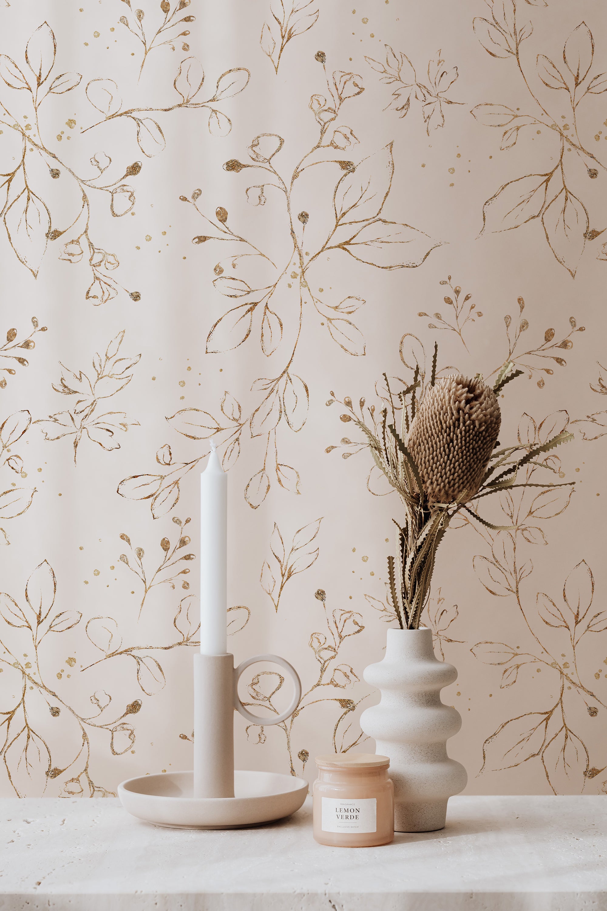 An elegant and sophisticated interior decor featuring Gold Leaves Wallpaper with hand-painted gold foliage patterns on a creamy background, complemented by a chic ceramic vase and candle holder.