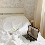 Cozy bedroom corner with Dainty Floral Line Wallpaper, showcasing a laptop on a bed with white linens and fresh flowers