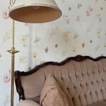 A classic living room corner, graced with the Pink Watercolour Floral Wallpaper featuring delicate hand-painted flowers in soft pink hues. The wallpaper adds a romantic touch behind a vintage tufted sofa, complemented by an elegant brass floor lamp that casts a warm glow, enhancing the room's inviting ambiance.