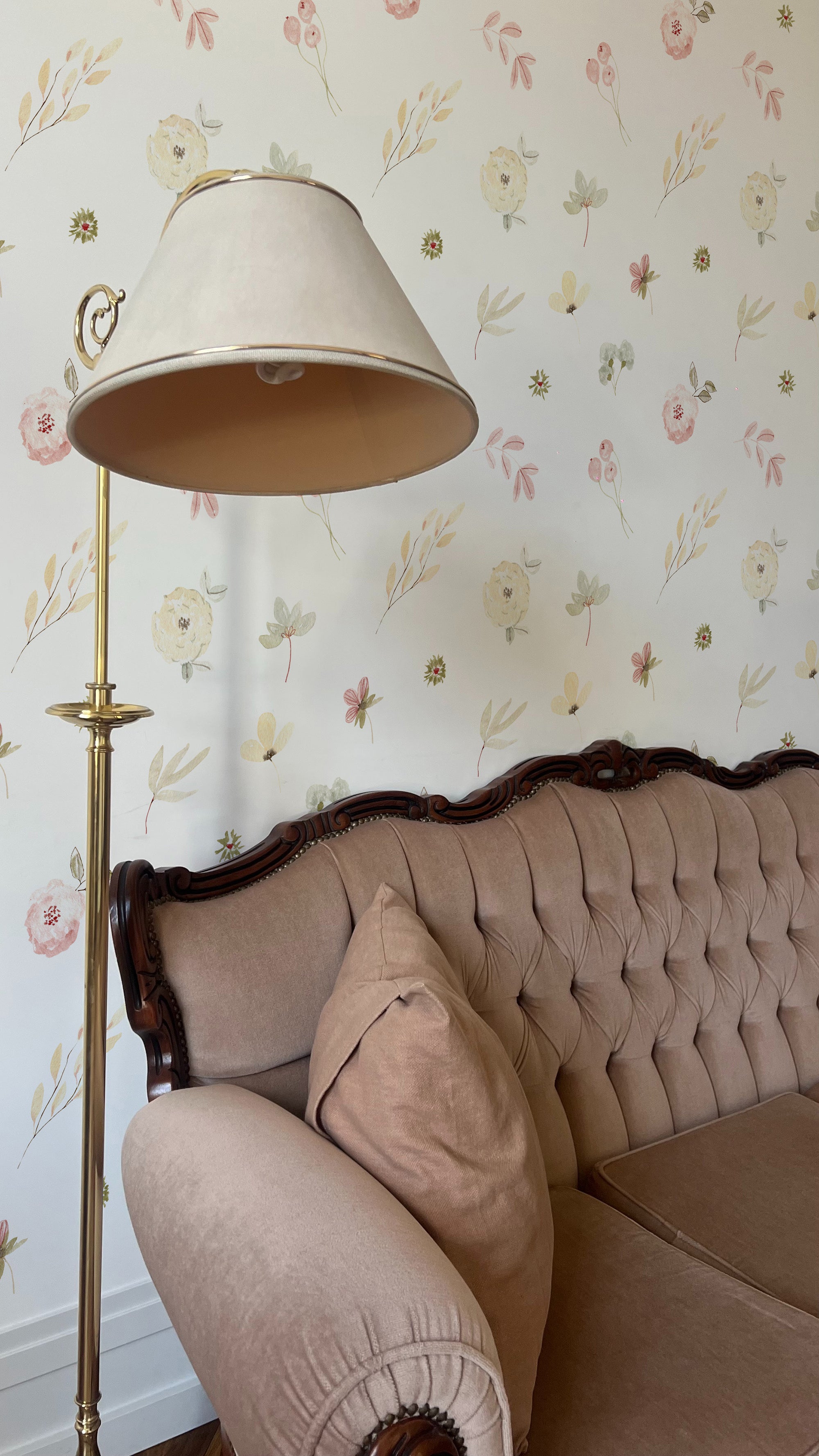 A classic living room corner, graced with the Pink Watercolour Floral Wallpaper featuring delicate hand-painted flowers in soft pink hues. The wallpaper adds a romantic touch behind a vintage tufted sofa, complemented by an elegant brass floor lamp that casts a warm glow, enhancing the room's inviting ambiance.