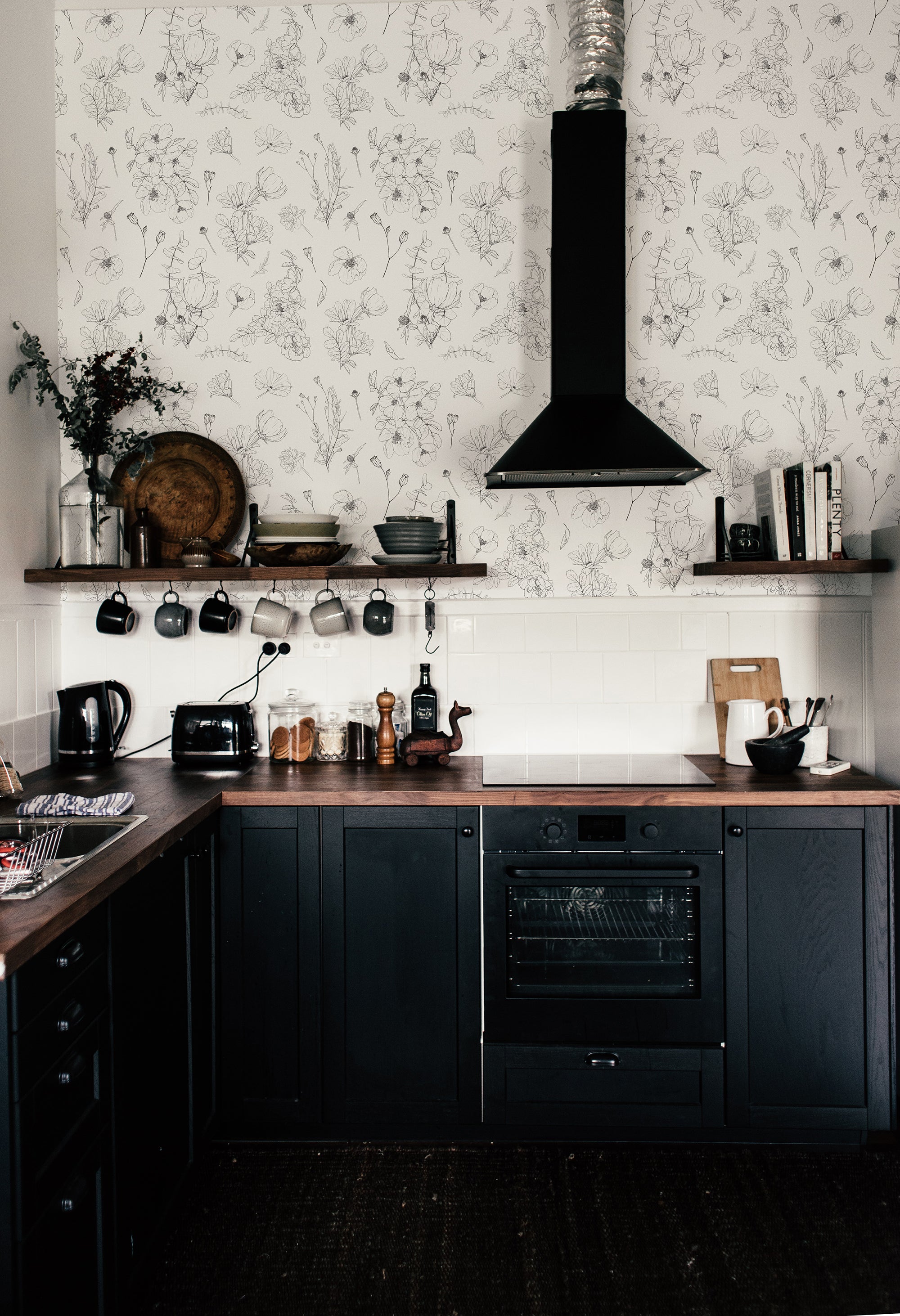 A stylish kitchen interior with dark wooden cabinets and a matching countertop, featuring a white subway tile backsplash and a wall adorned with 'Wildflower Sketch Wallpaper,' which displays a monochromatic floral pattern of various wildflowers and leaves in a hand-drawn style.