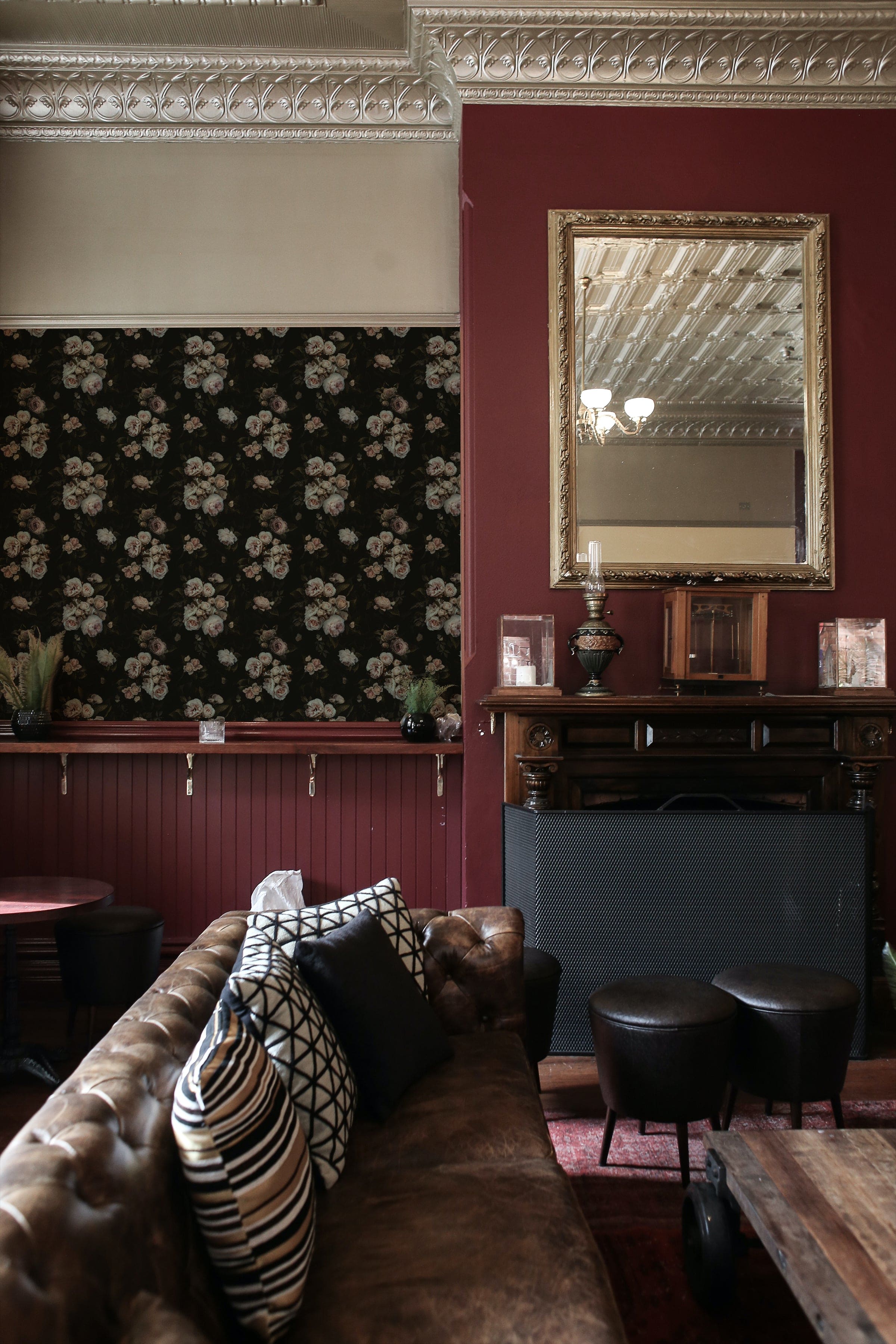 A stylish interior setting featuring the Dark Rose Wallpaper used as a dramatic accent wall in a lounge area. The deep tones of the roses complement the vintage leather sofa and classic decor elements, enhancing the room's sophisticated, dark aesthetic.