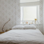 A bright and peaceful bedroom featuring the Tranquil Bloom Wallpaper II. The room is bathed in natural light from a window, highlighting the wallpaper's delicate floral design in soft, muted colors that evoke a feeling of serenity. A white metal bed is neatly made with a beige linen duvet, and a white tray with a small plant is placed by the window, enhancing the room’s tranquil vibe.