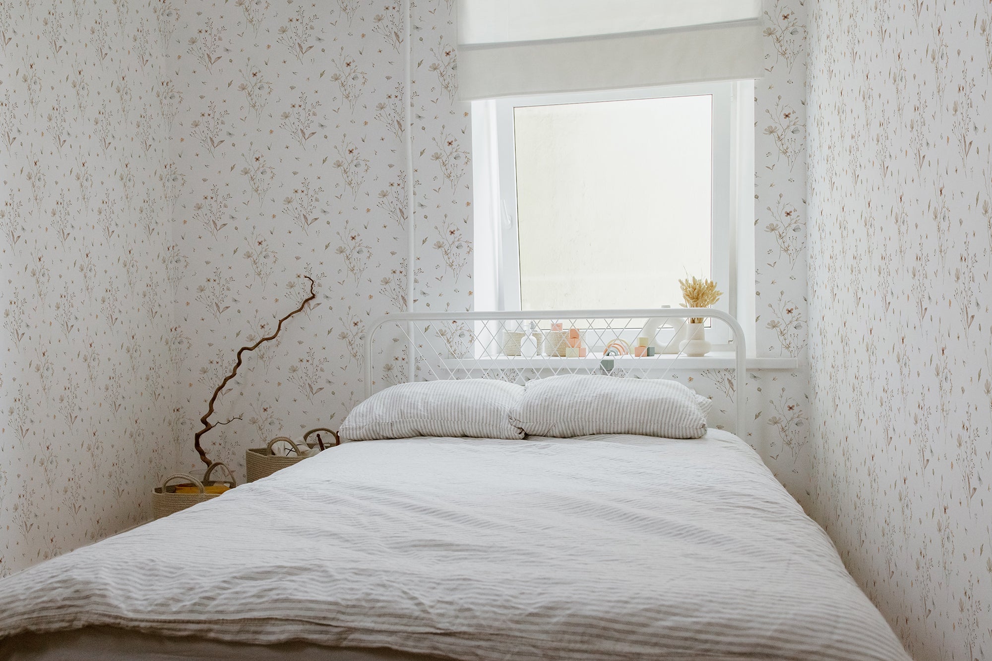 A bright and peaceful bedroom featuring the Tranquil Bloom Wallpaper II. The room is bathed in natural light from a window, highlighting the wallpaper's delicate floral design in soft, muted colors that evoke a feeling of serenity. A white metal bed is neatly made with a beige linen duvet, and a white tray with a small plant is placed by the window, enhancing the room’s tranquil vibe.