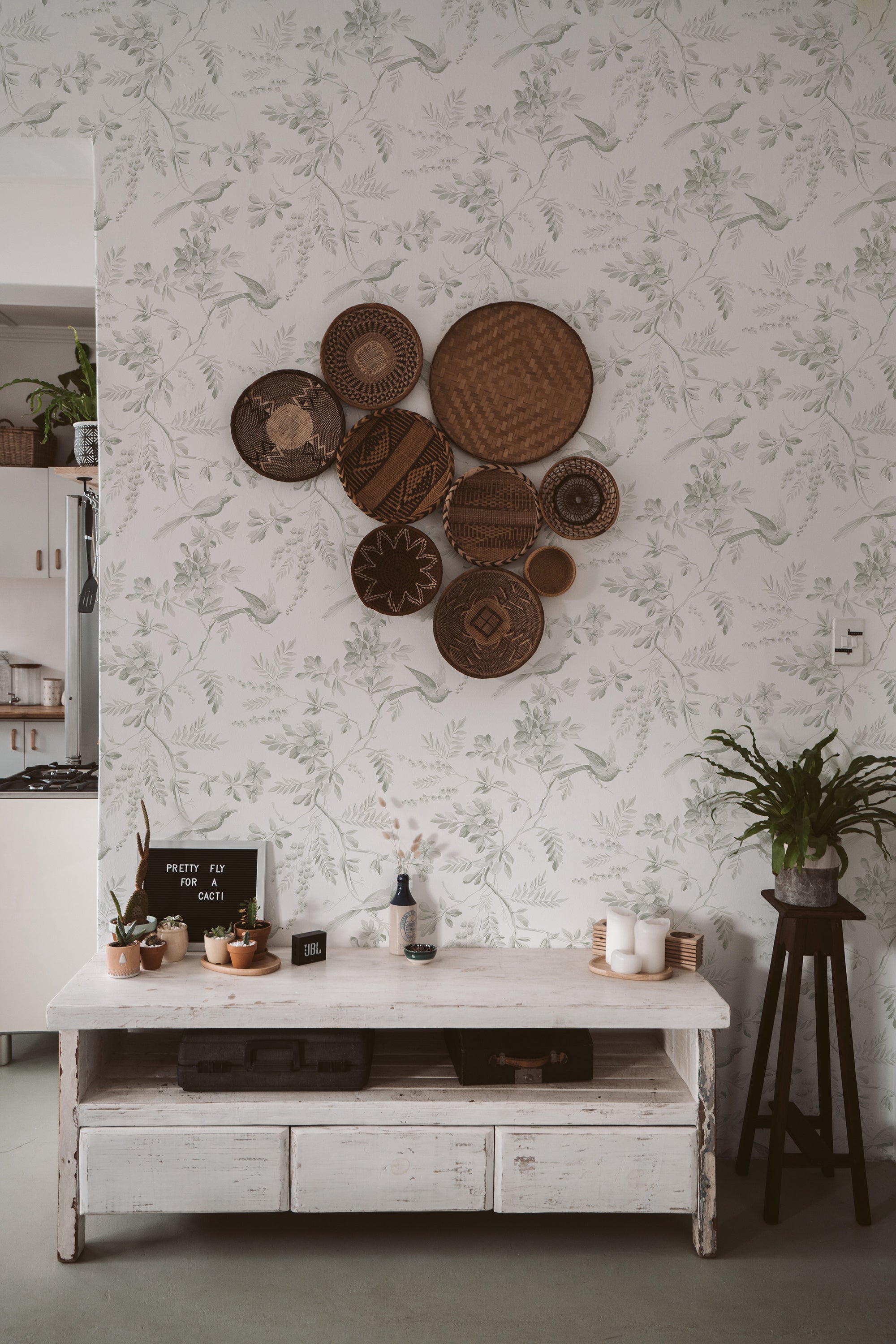 An elegantly styled room with 'Blossoming Birds Wallpaper' on the wall, featuring intricate illustrations of birds and flora in shades of green, with a collection of woven baskets as a creative wall decoration, and a rustic white console table with cacti.
