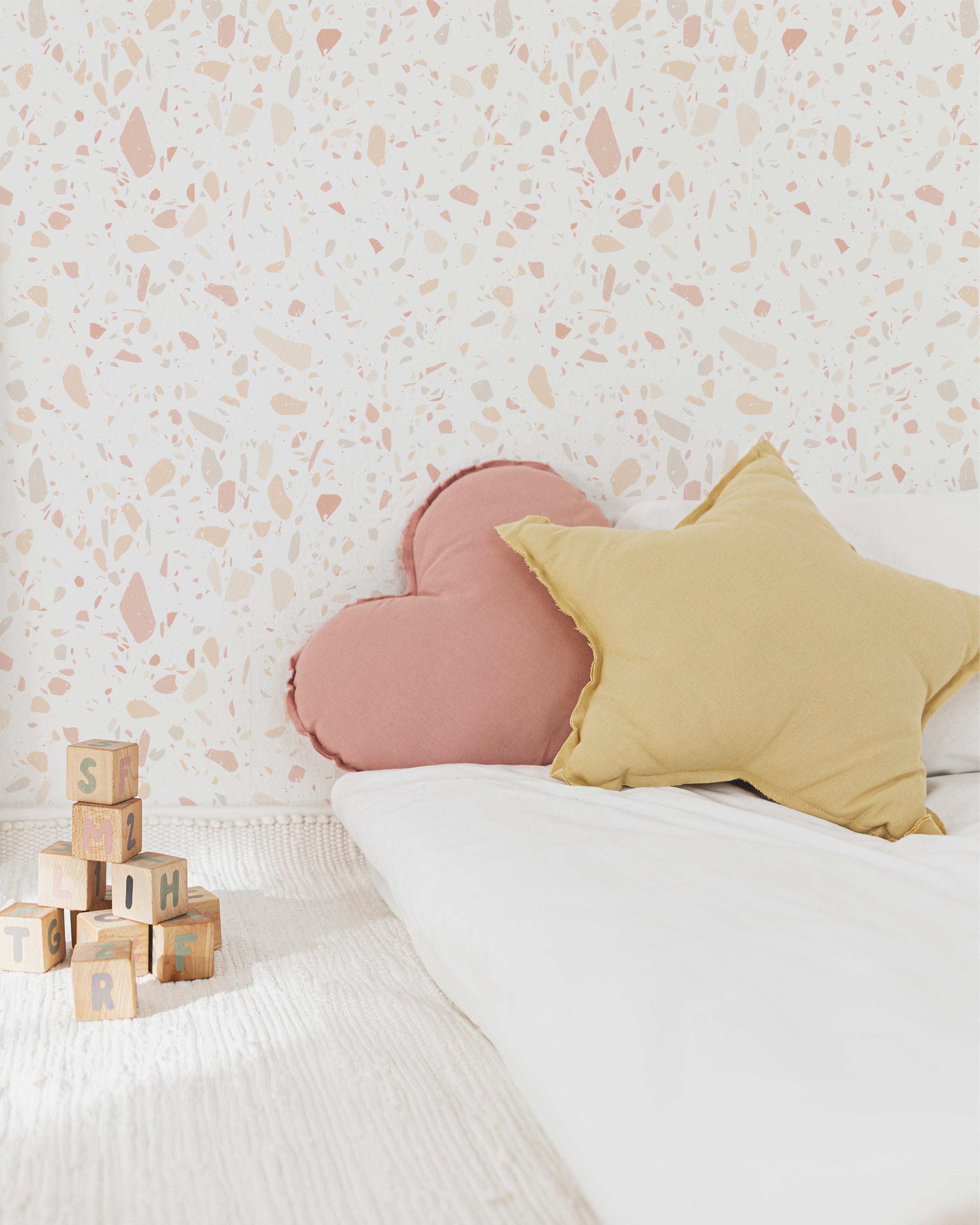 A nursery room featuring the Pink Terrazzo Wallpaper which adds a subtle and modern backdrop. The wallpaper complements the room's light, pastel-themed decor including soft pillows and children's toys, creating a cheerful and contemporary space.