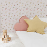 A child's bedroom adorned with Pink Watercolour Floral Wallpaper II. The wall is decorated with a pattern of soft pink flowers and green leaves. The scene includes a bed with pink and mustard yellow pillows and wooden alphabet blocks scattered on a white textured rug