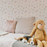 A cozy bedroom corner featuring Pink Watercolour Floral Wallpaper II with a pattern of delicate pink and beige flowers scattered over a pale background. A large plush monkey and a stuffed dog sit on a textured gray crochet blanket, with decorative pillows adding a touch of elegance.