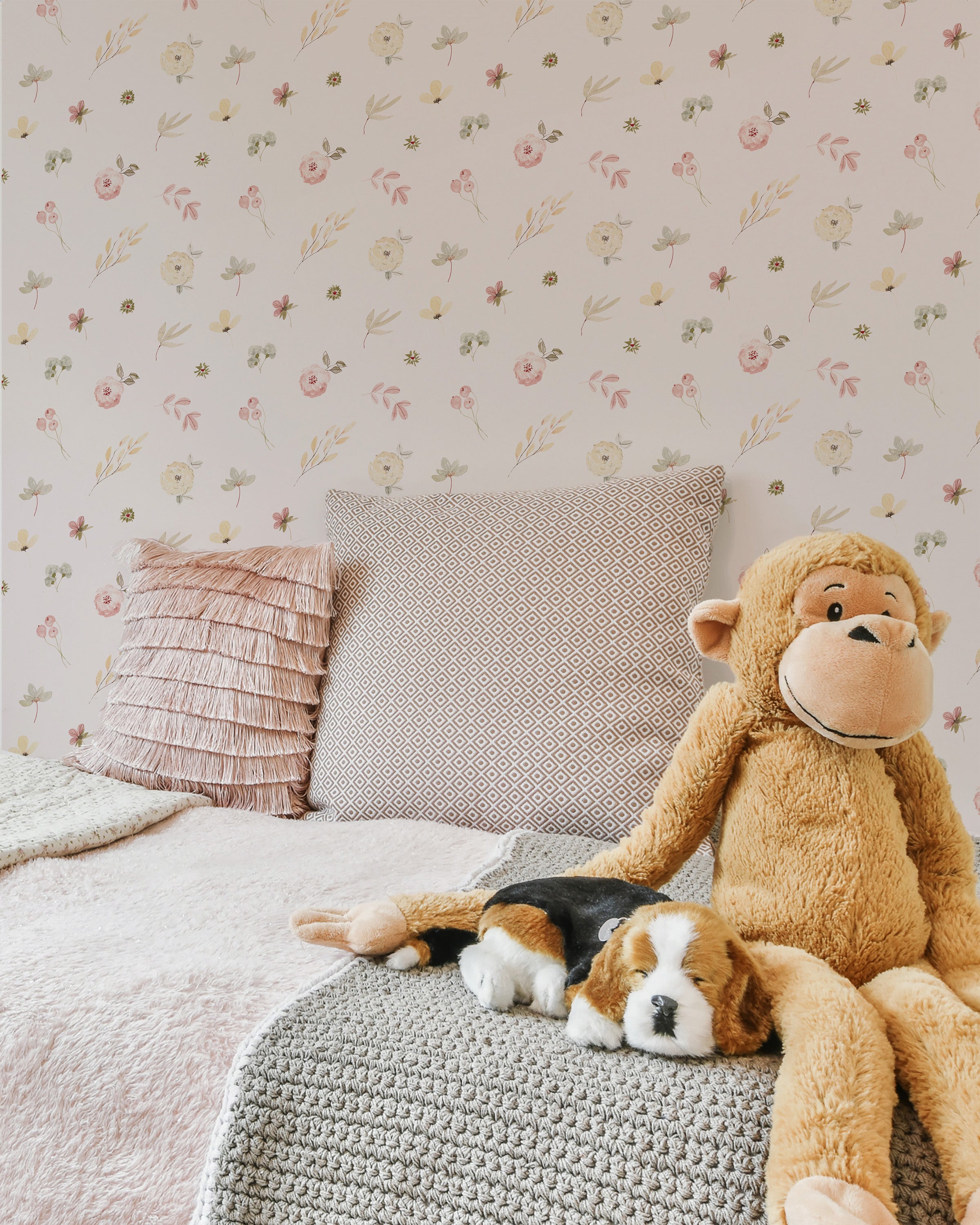 A cozy bedroom corner featuring Pink Watercolour Floral Wallpaper II with a pattern of delicate pink and beige flowers scattered over a pale background. A large plush monkey and a stuffed dog sit on a textured gray crochet blanket, with decorative pillows adding a touch of elegance.