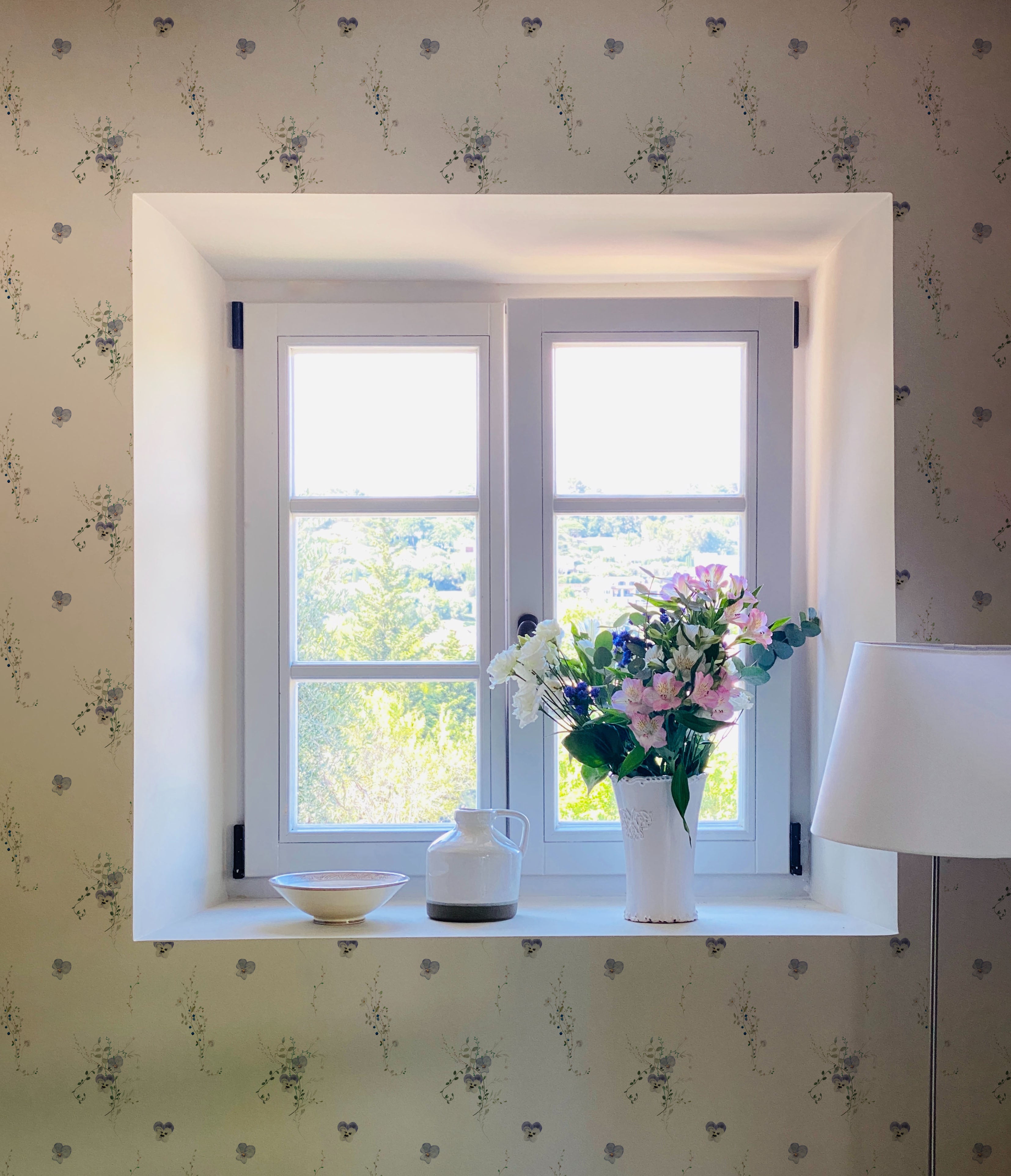 The Delicate Blue Wildflower Wallpaper accents a bright window area, bringing the gentle beauty of a wildflower meadow indoors. A vase with fresh flowers on the windowsill echoes the wallpaper's floral motif.