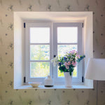 The Delicate Blue Wildflower Wallpaper accents a bright window area, bringing the gentle beauty of a wildflower meadow indoors. A vase with fresh flowers on the windowsill echoes the wallpaper's floral motif.