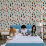 Children's room decorated with a vibrant 'Tidal Sail Wallpaper' featuring a colorful array of origami sailboats on a creamy background. Three young children are engaged in activities on a blue bed, adding a lively atmosphere to the room.