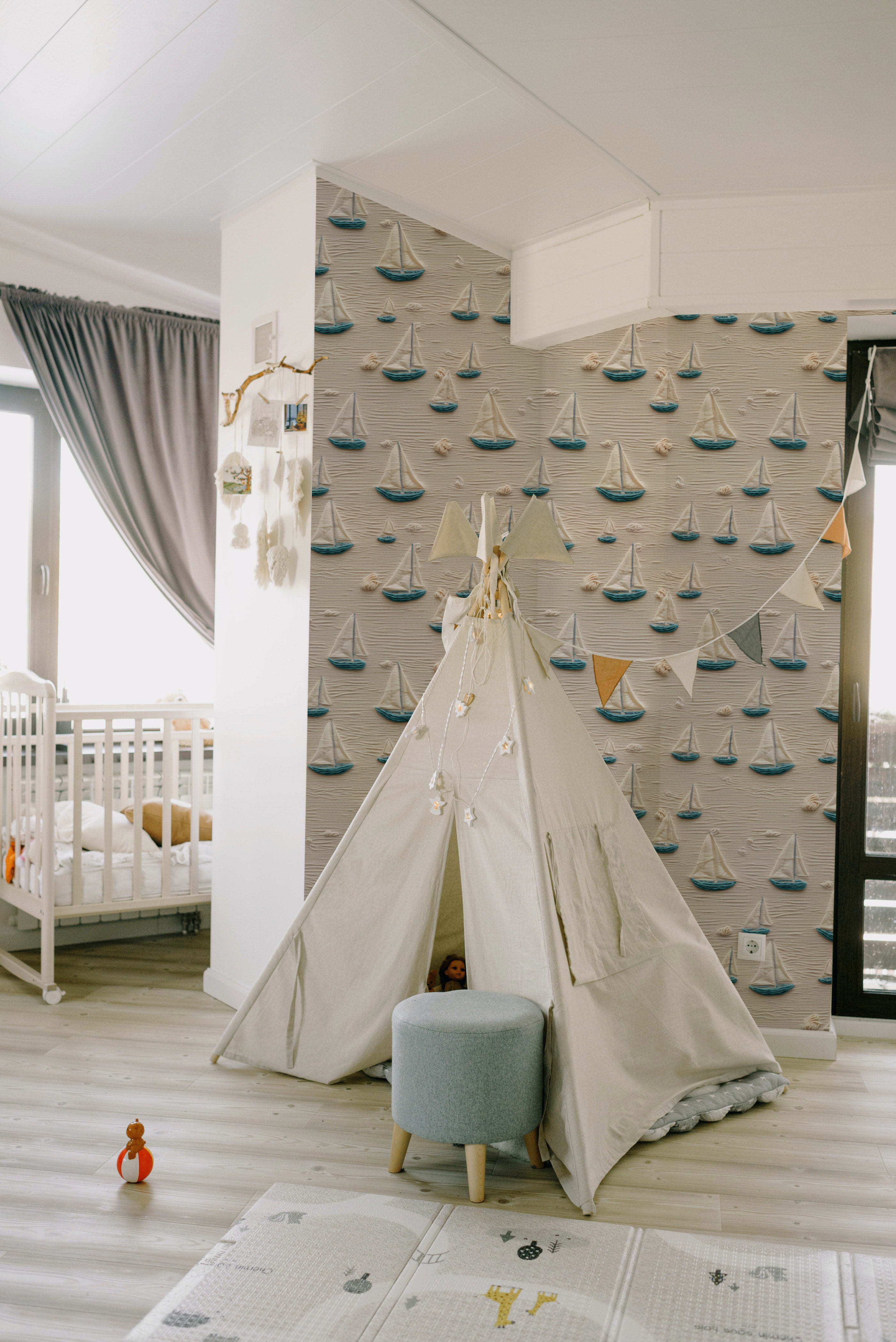 A child's play area with a tent set up against a wall covered in Portside Sail Wallpaper, which features an array of textured sailboats on a sandy beige background, evoking the calm of a seaside marina.