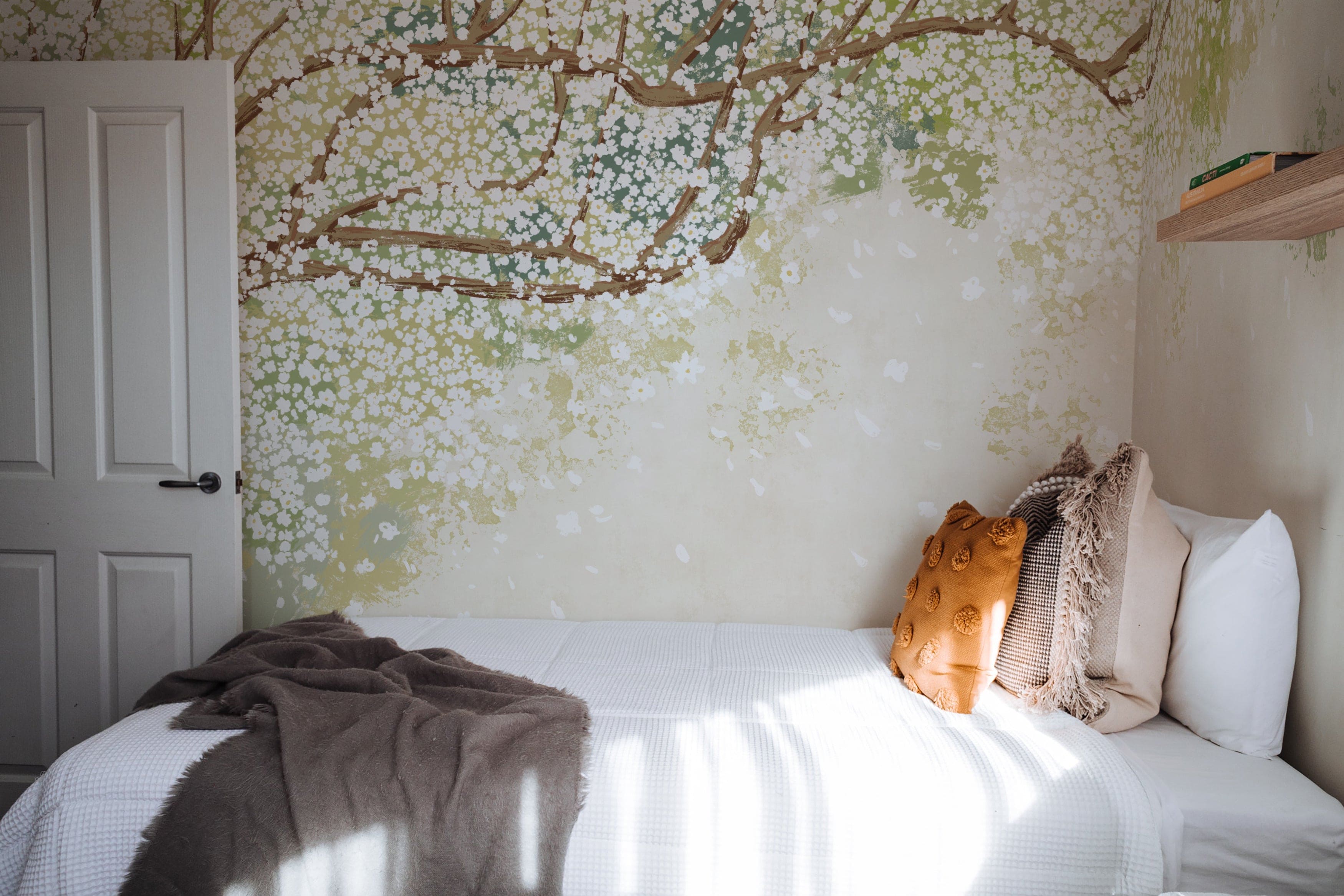 A cozy bedroom with a bed and a few pillows placed against a wall adorned with the Sakura Serenade mural wallpaper. The mural features branches of cherry blossoms with white flowers on a soft green and beige backdrop, adding a calming and natural touch to the room.