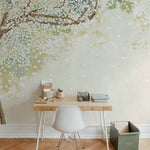 A serene home office setup with a wooden desk and white chair against a wall covered in a Sakura Serenade mural wallpaper. The wallpaper features delicate cherry blossom branches with white flowers set against a soft green and beige background, creating a peaceful and inspiring work environment.
