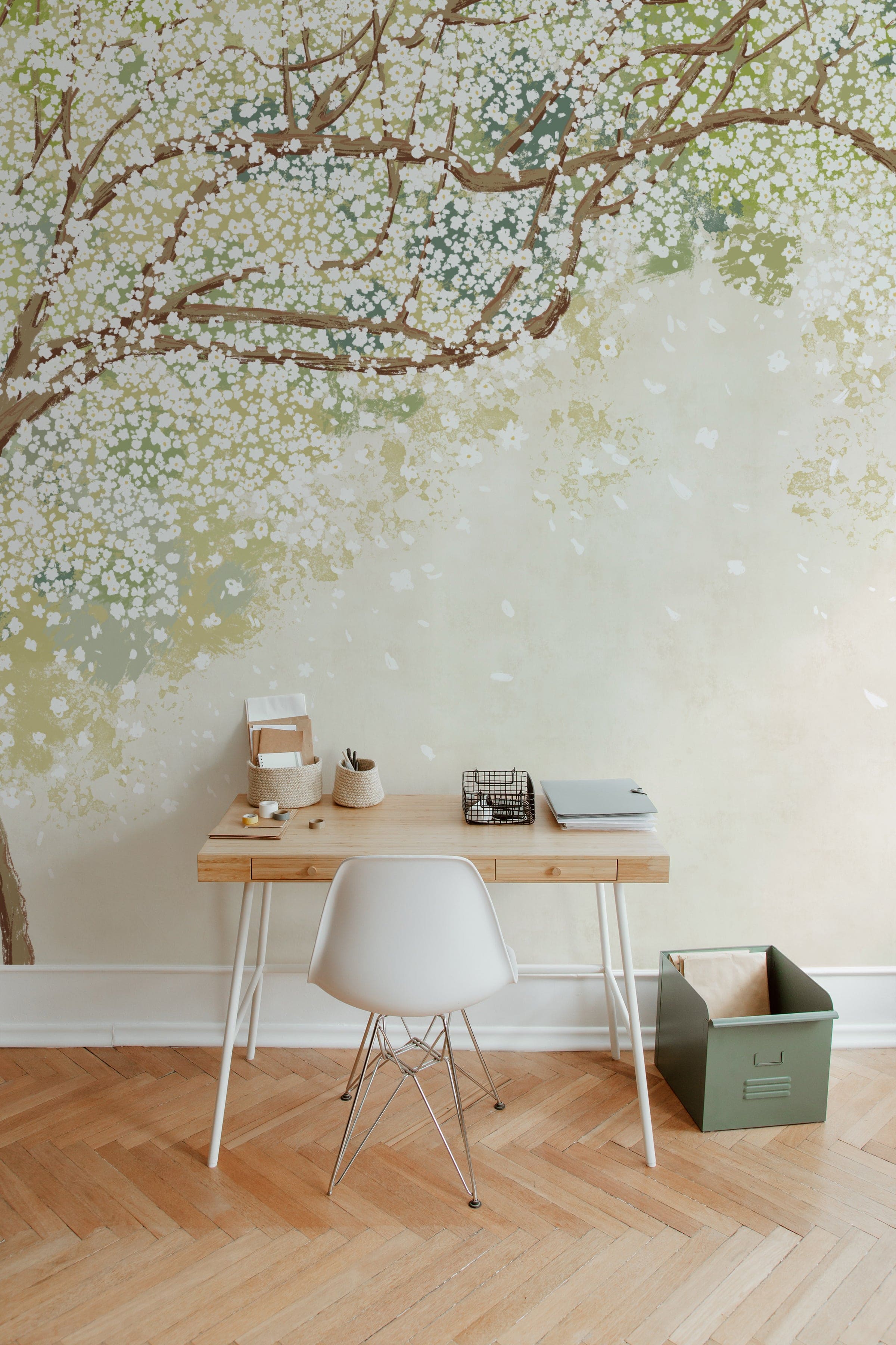 A serene home office setup with a wooden desk and white chair against a wall covered in a Sakura Serenade mural wallpaper. The wallpaper features delicate cherry blossom branches with white flowers set against a soft green and beige background, creating a peaceful and inspiring work environment.