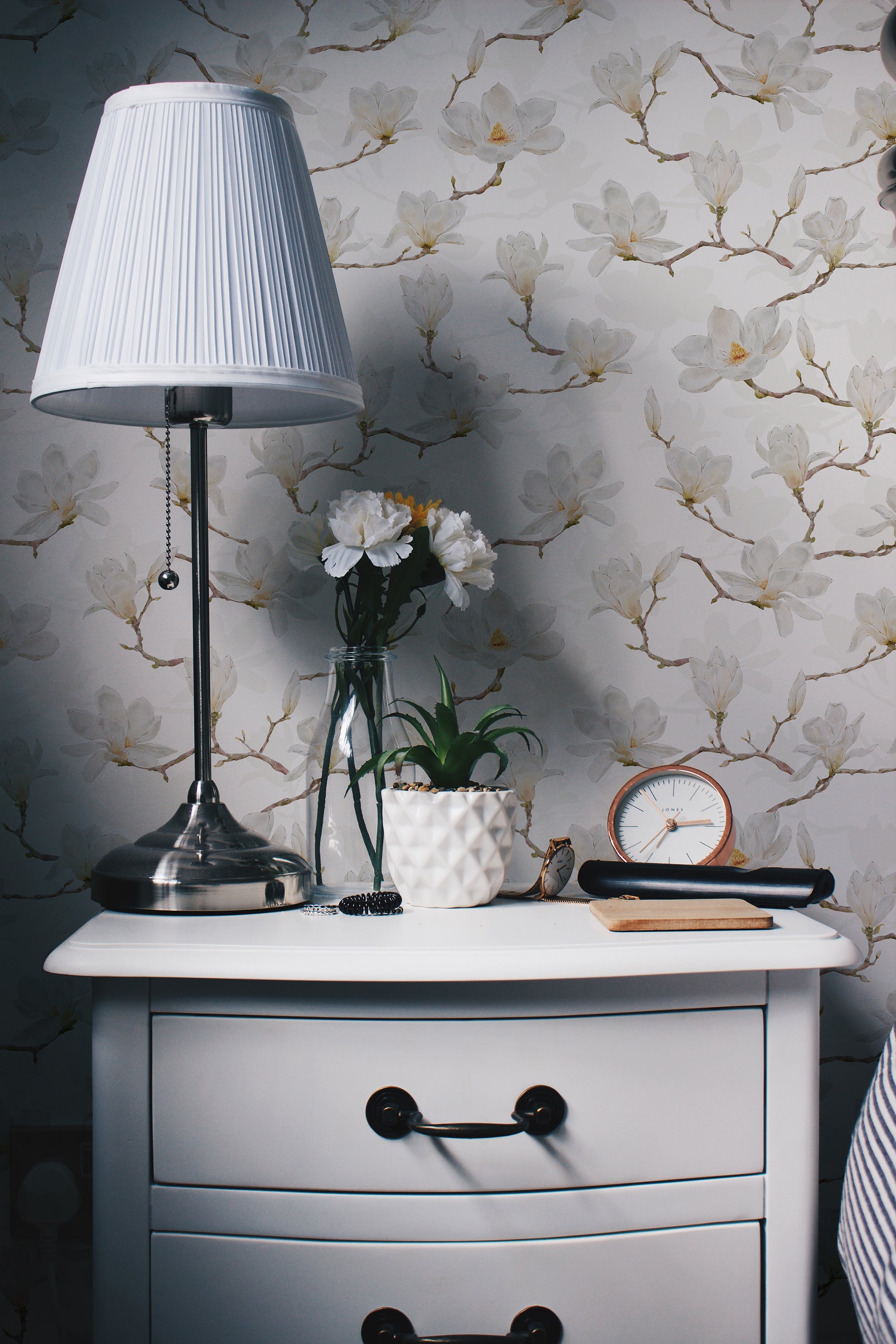 A cozy bedside table setup featuring a metallic lamp with a pleated white lampshade, a glass vase with white flowers, a small potted plant in a white geometric pot, a round clock, and some accessories. The background showcases a beautiful white watercolour magnolia wallpaper.