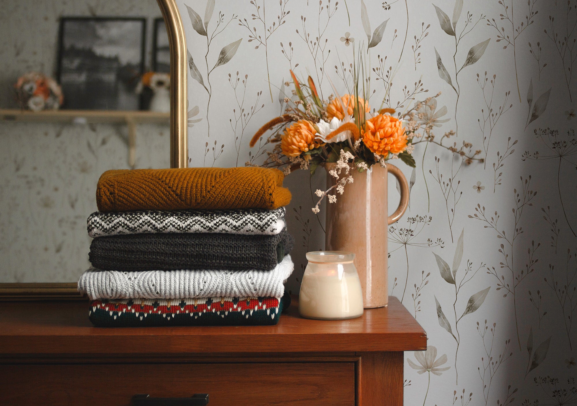 A stylish living space accented with Tranquil Bloom Wallpaper, where the muted floral pattern adds an elegant backdrop to the folded autumnal-colored knitwear and ceramic vase with vibrant flowers.