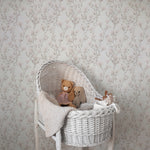 A cozy nursery corner showcasing the Vintage Floral Reverie Wallpaper as a gentle and refined backdrop, with soft pink blooms and gray foliage complementing the white wicker bassinet and plush teddy bears.