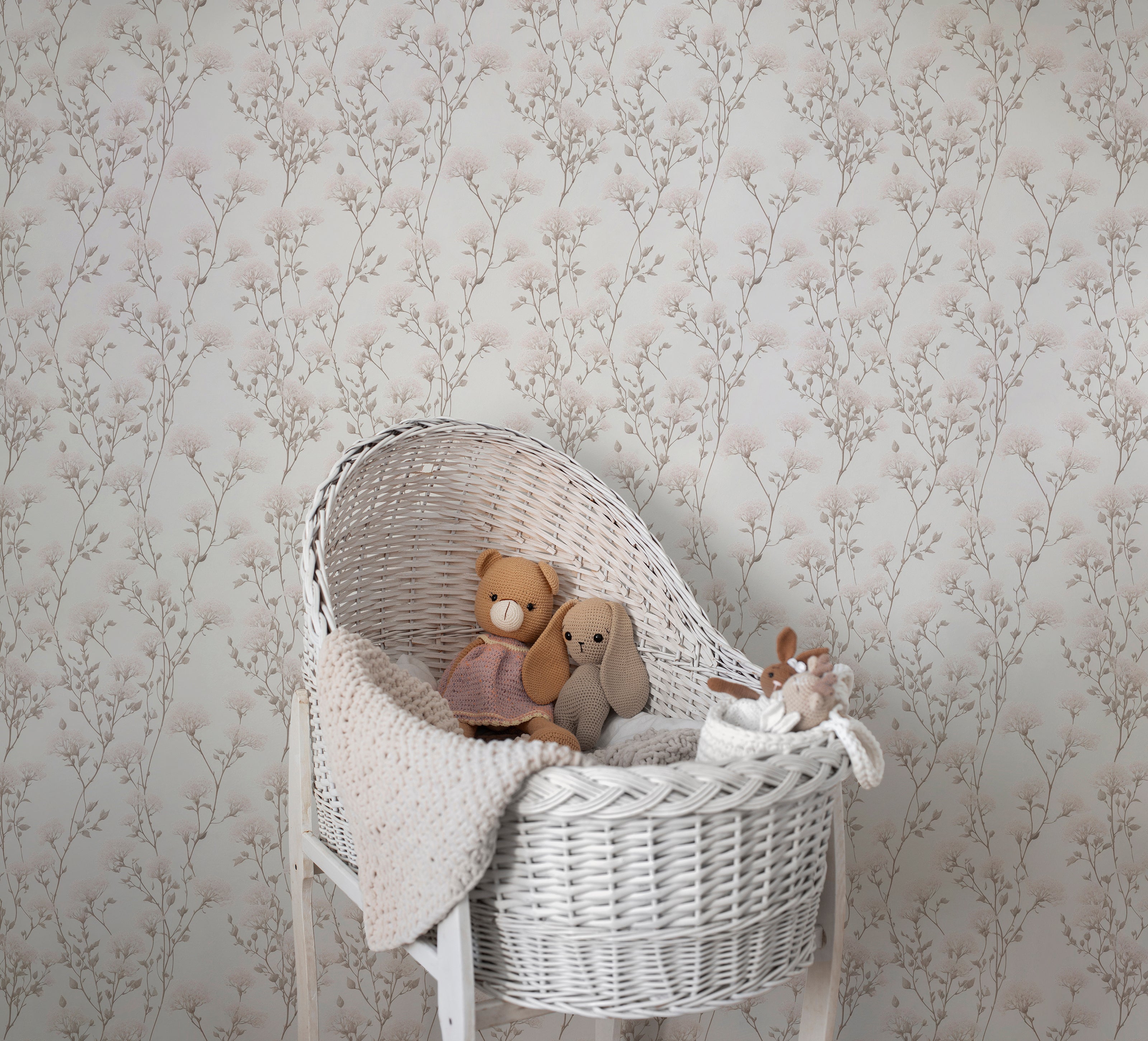 A cozy nursery corner showcasing the Vintage Floral Reverie Wallpaper as a gentle and refined backdrop, with soft pink blooms and gray foliage complementing the white wicker bassinet and plush teddy bears.