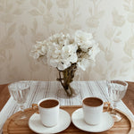 A simple yet stylish home coffee setup with the Cream Watercolour Rose Wallpaper in the background. The scene includes a wooden tray on a striped tablecloth holding two cups of coffee and a vase with white flowers, creating a serene morning atmosphere.