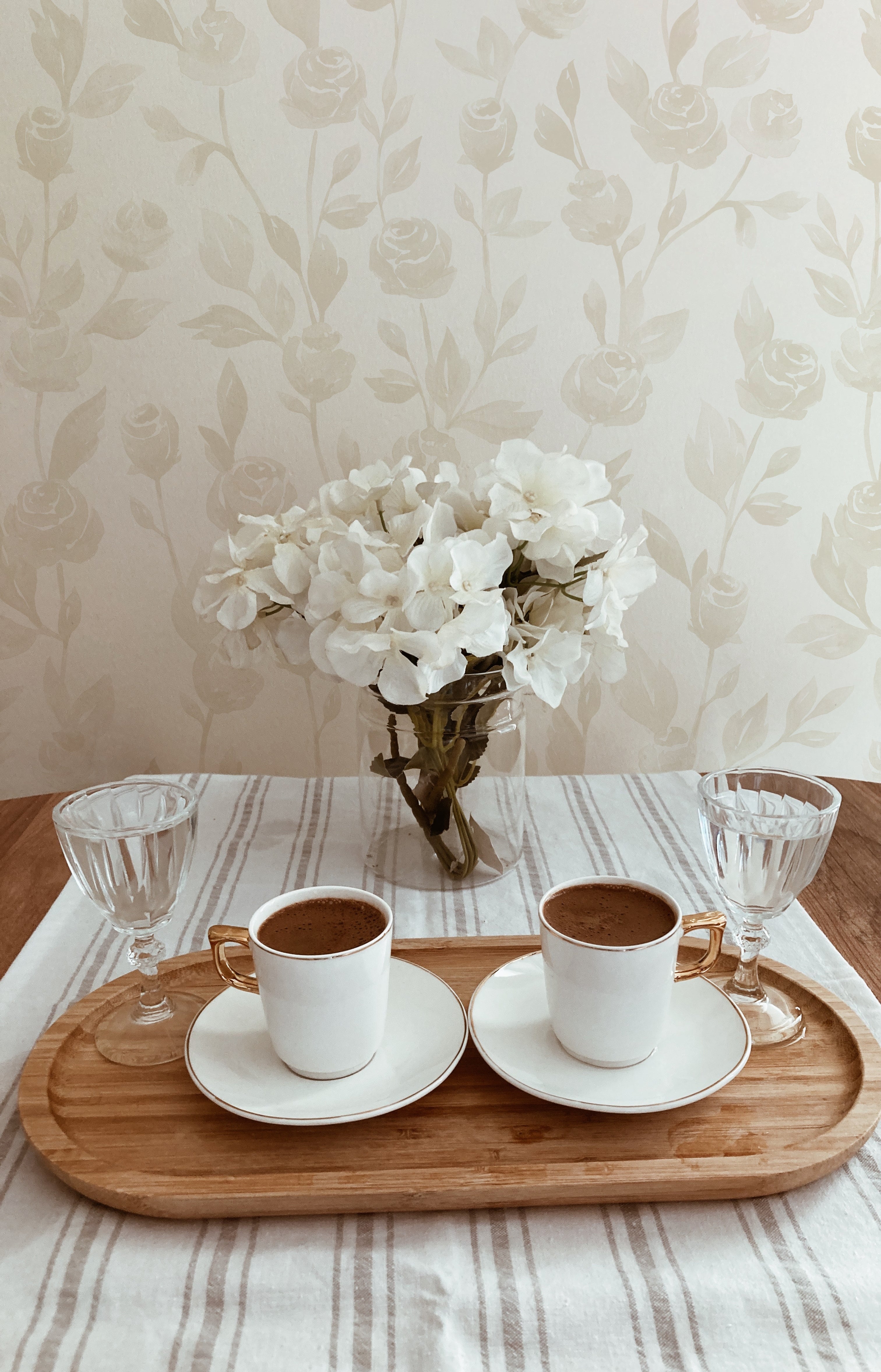 A simple yet stylish home coffee setup with the Cream Watercolour Rose Wallpaper in the background. The scene includes a wooden tray on a striped tablecloth holding two cups of coffee and a vase with white flowers, creating a serene morning atmosphere.