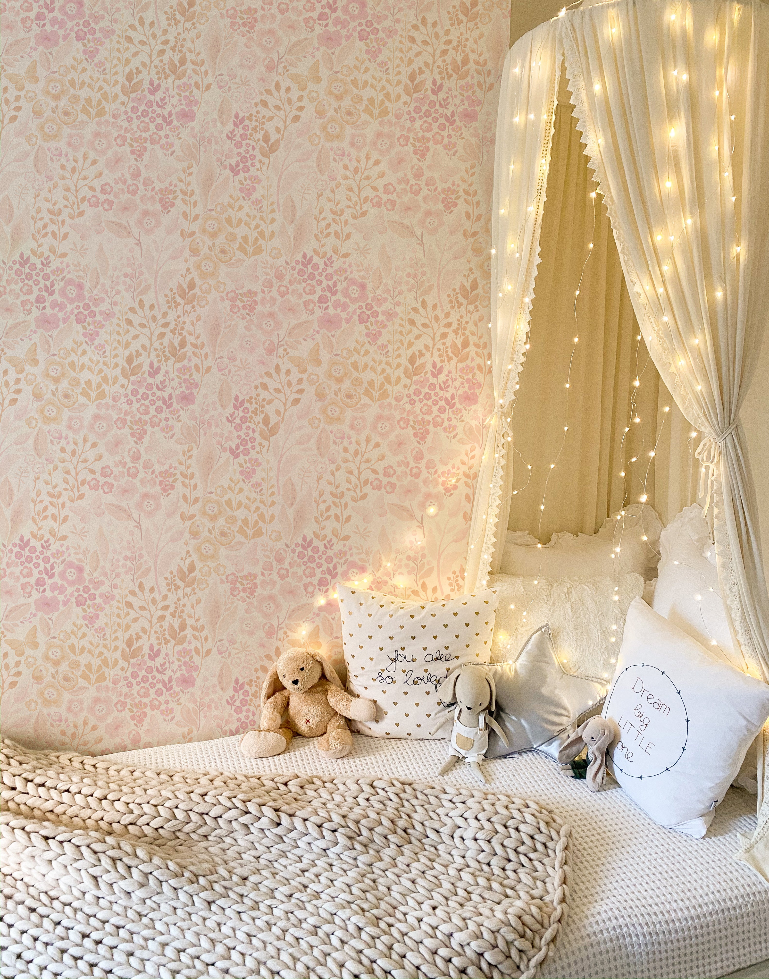 A whimsical child's bedroom is wrapped in the warmth of the Pretty Petals Wallpaper - 25", featuring a delicate floral print. The room is enchanted by a canopy of fairy lights, plush pillows, and cuddly toys on the bed, offering a magical and peaceful retreat for a child.