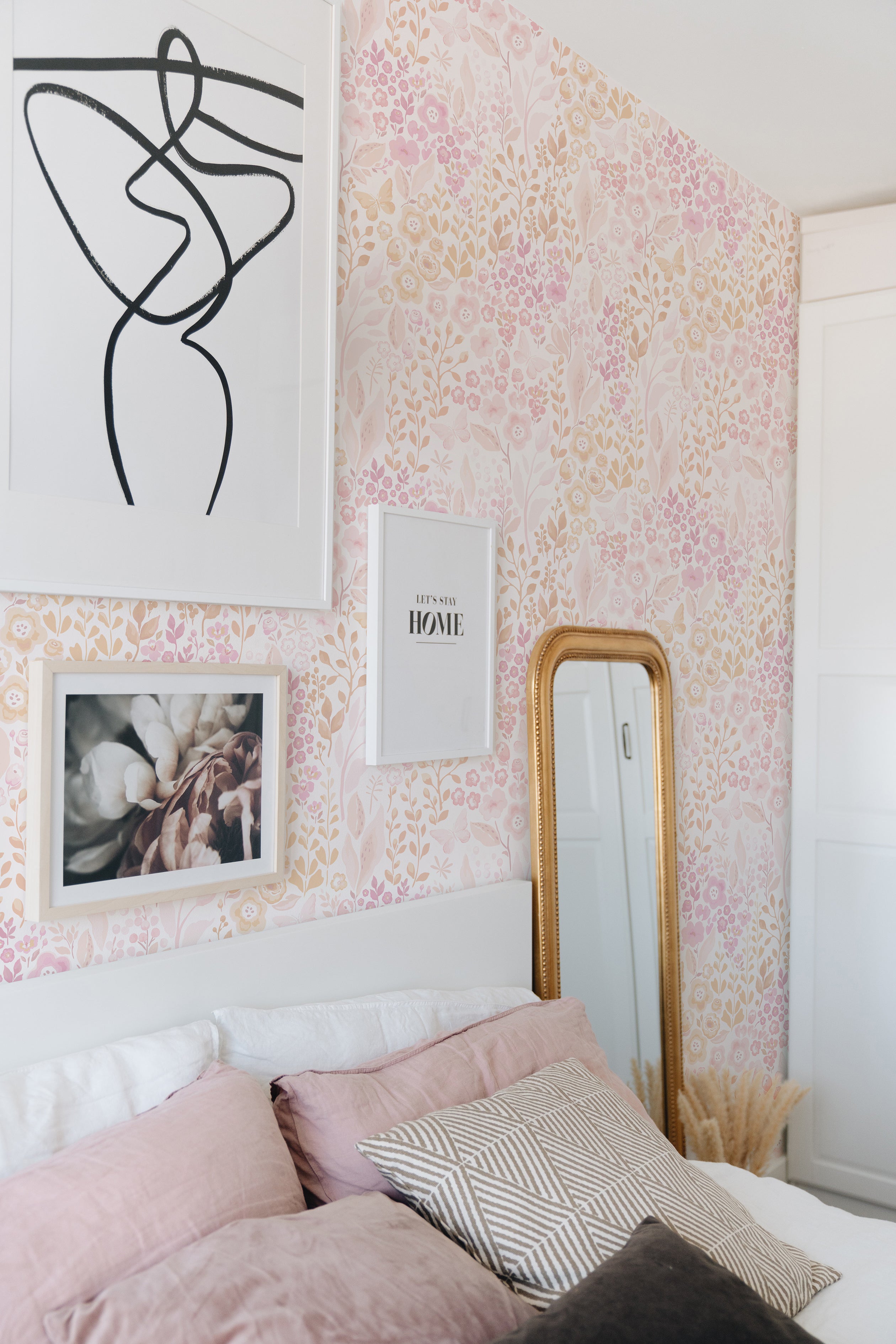 A cozy corner of a bedroom exudes charm with the Pretty Petals Wallpaper - 25". The wall, covered in soft pink and beige florals, provides a delicate backdrop to a classic bed with pink linens, framed art pieces, and an ornate gold mirror, creating a warm and stylish sleeping area.