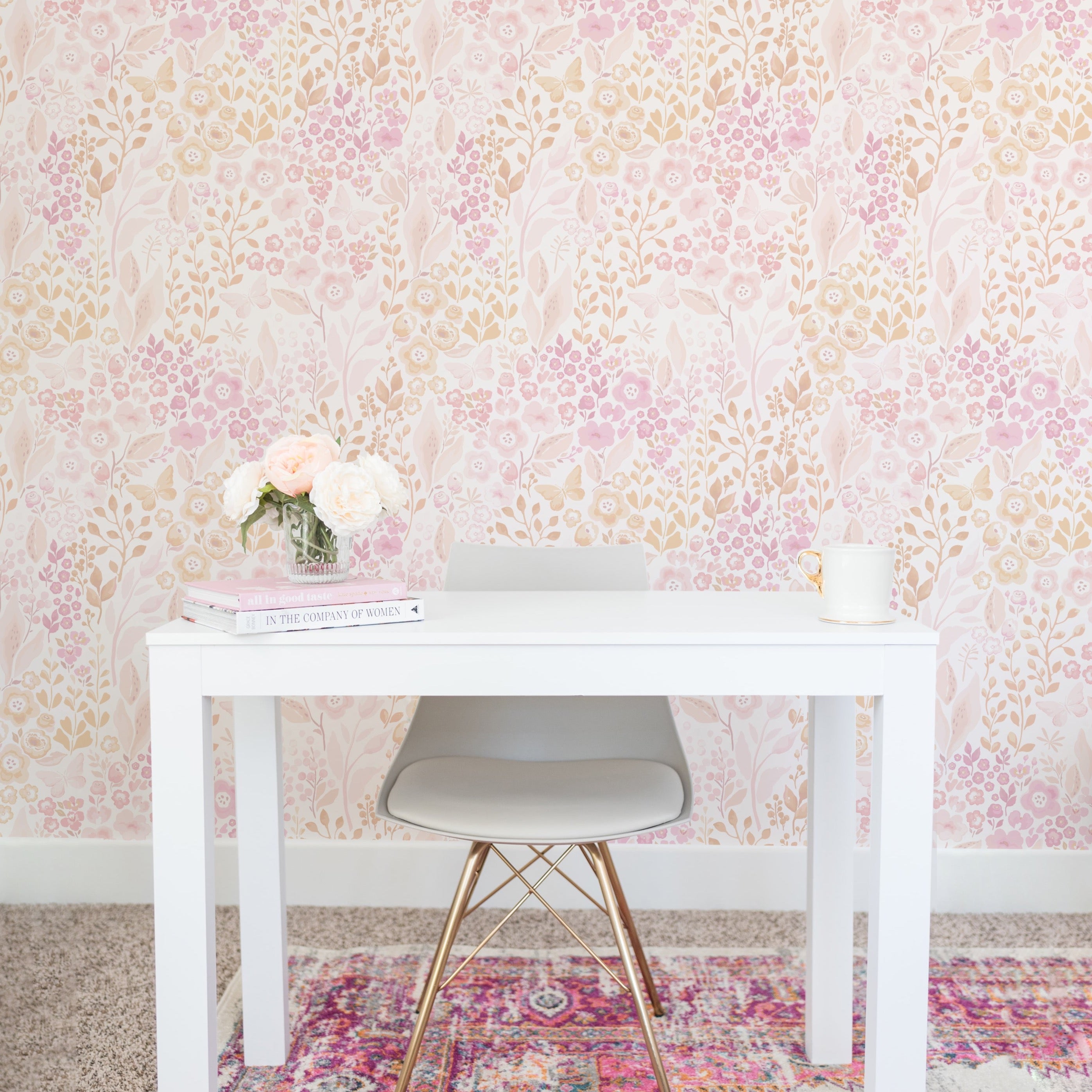 A bright and inviting workspace featuring the Pretty Petals Wallpaper - 25" adorned with blush pink and beige floral patterns. The wallpaper adds a romantic touch to the space, complementing a sleek white desk, a modern gray chair with golden legs, and a vibrant multicolored rug beneath.
