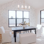 A bright and airy dining room with large windows, a black dining table, white upholstered chairs, and a pendant chandelier. The walls are decorated with white wallpaper showcasing an abstract black floral pattern, enhancing the room's elegance.