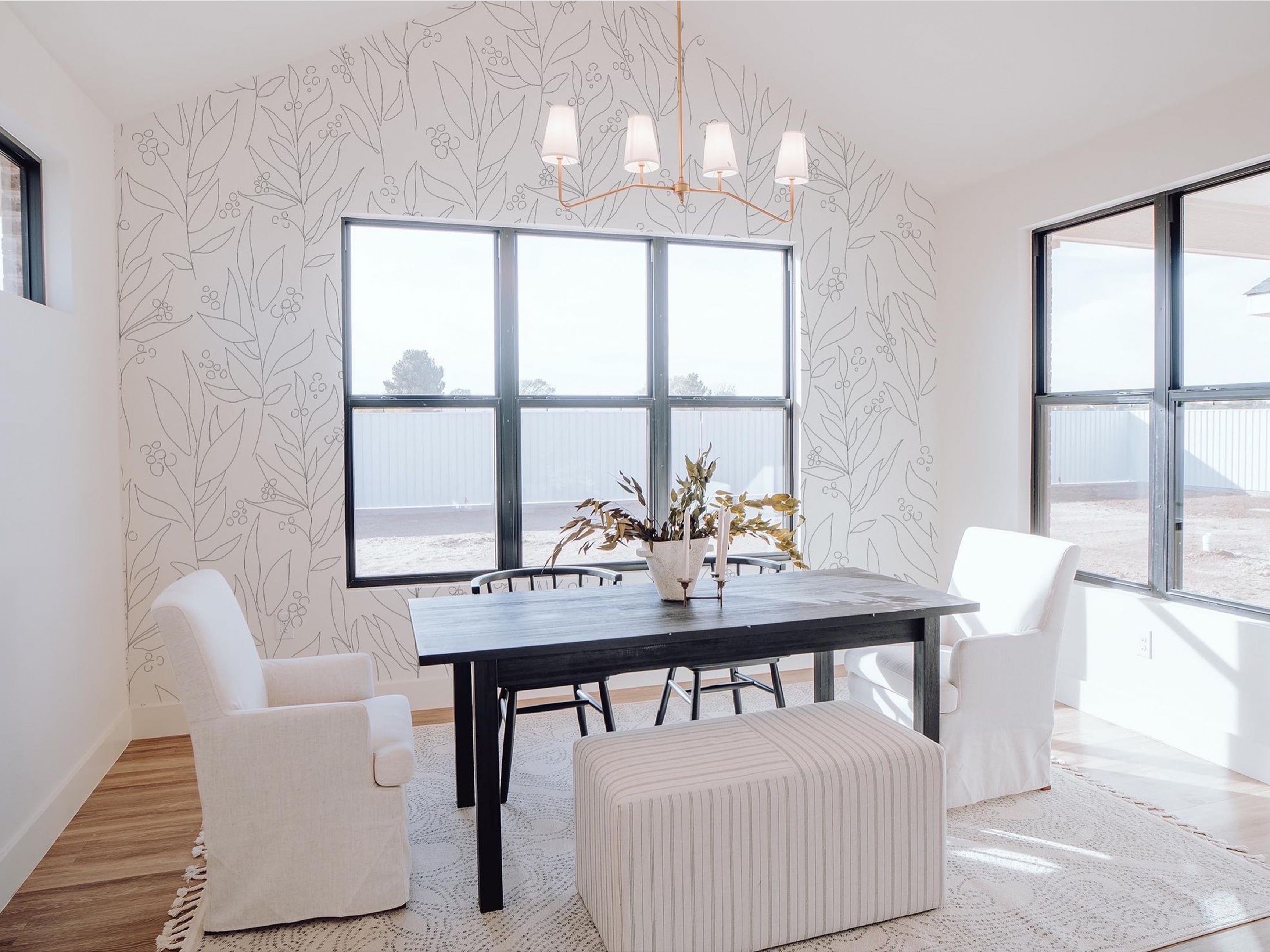 A bright and airy dining room with large windows, a black dining table, white upholstered chairs, and a pendant chandelier. The walls are decorated with white wallpaper showcasing an abstract black floral pattern, enhancing the room's elegance.