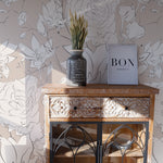 A stylish vignette featuring an ornate wooden cabinet against a wall adorned with the Floral Beauty Wallpaper. The wallpaper showcases large-scale, monochrome floral outlines in shades of white and gray on a taupe background. A decorative vase with dried flowers and a book labeled "BON APPETIT" add a rustic charm to the setting.