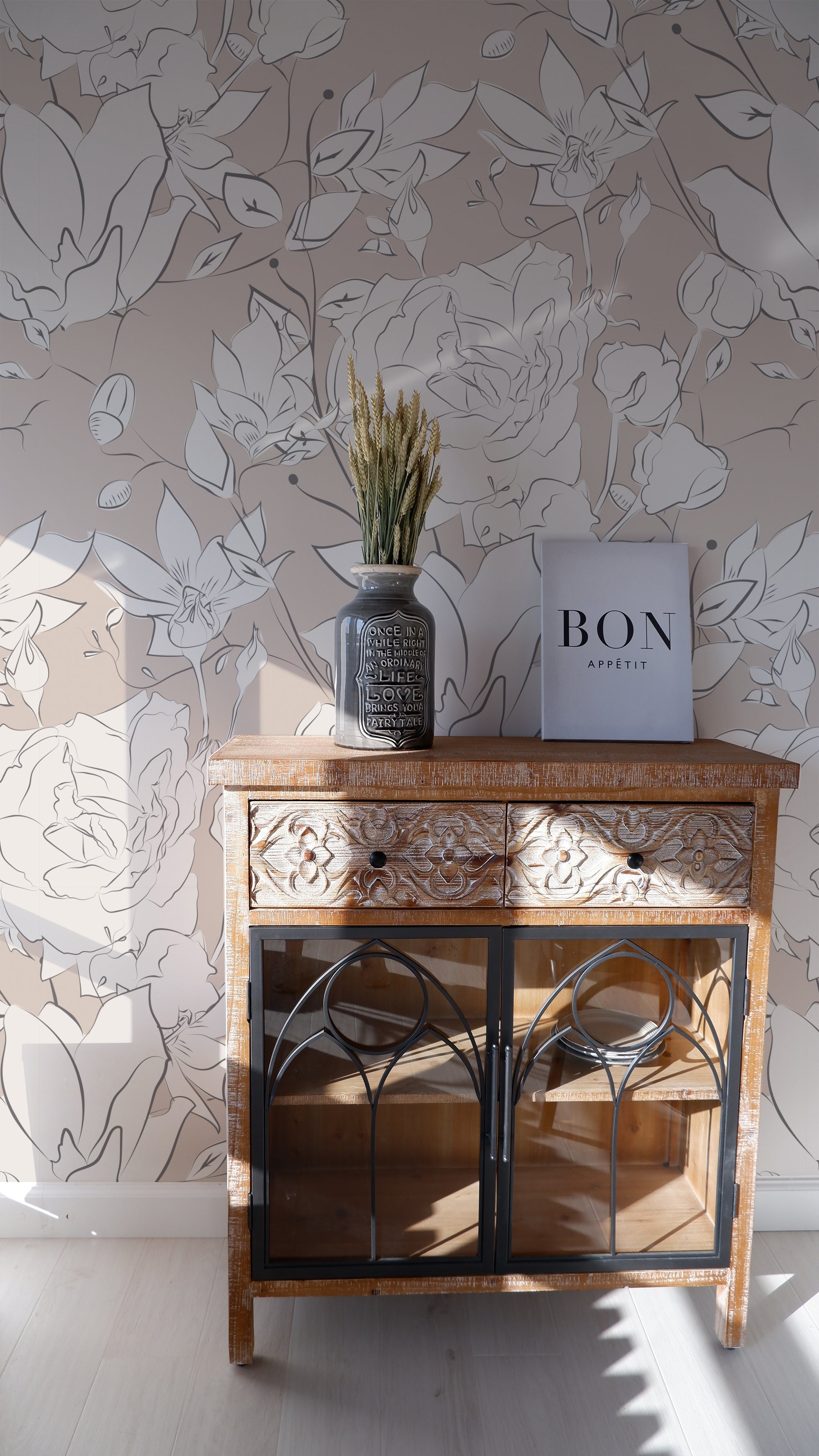 A stylish vignette featuring an ornate wooden cabinet against a wall adorned with the Floral Beauty Wallpaper. The wallpaper showcases large-scale, monochrome floral outlines in shades of white and gray on a taupe background. A decorative vase with dried flowers and a book labeled "BON APPETIT" add a rustic charm to the setting.