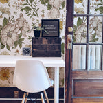 an image showcasing the "Floral Wallpaper - Sunny II" in an office space. The wallpaper is patterned with large, sunny floral illustrations in warm tones, giving the space a vibrant yet sophisticated feel. A modern white chair contrasts with the rustic wooden elements in the room, complementing the natural aesthetic of the wallpaper.