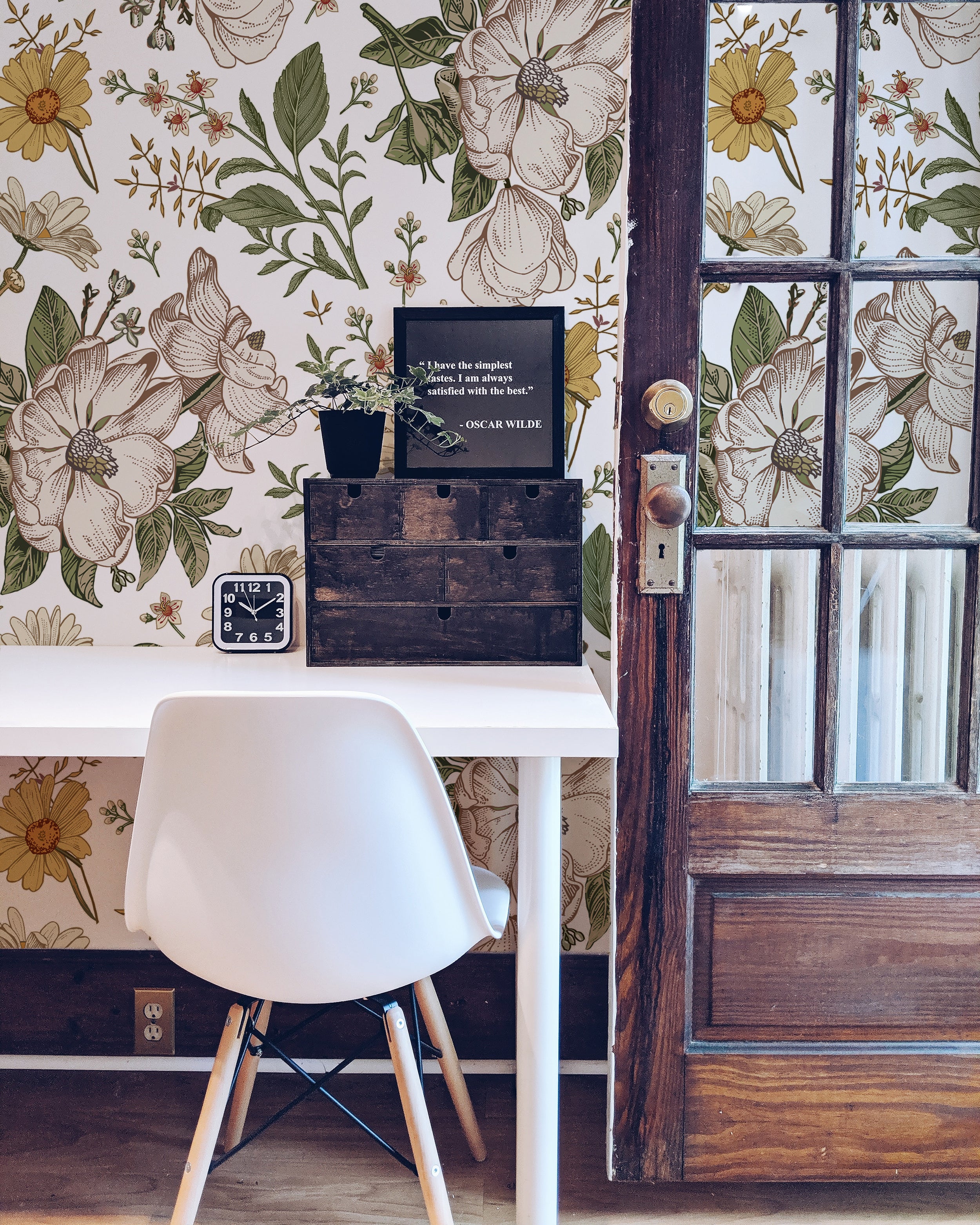 an image showcasing the "Floral Wallpaper - Sunny II" in an office space. The wallpaper is patterned with large, sunny floral illustrations in warm tones, giving the space a vibrant yet sophisticated feel. A modern white chair contrasts with the rustic wooden elements in the room, complementing the natural aesthetic of the wallpaper.
