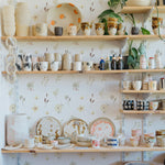 A lively and bright kitchen space with shelves of handcrafted pottery against the Sunny Floral Wallpaper, creating a homey and inviting atmosphere.