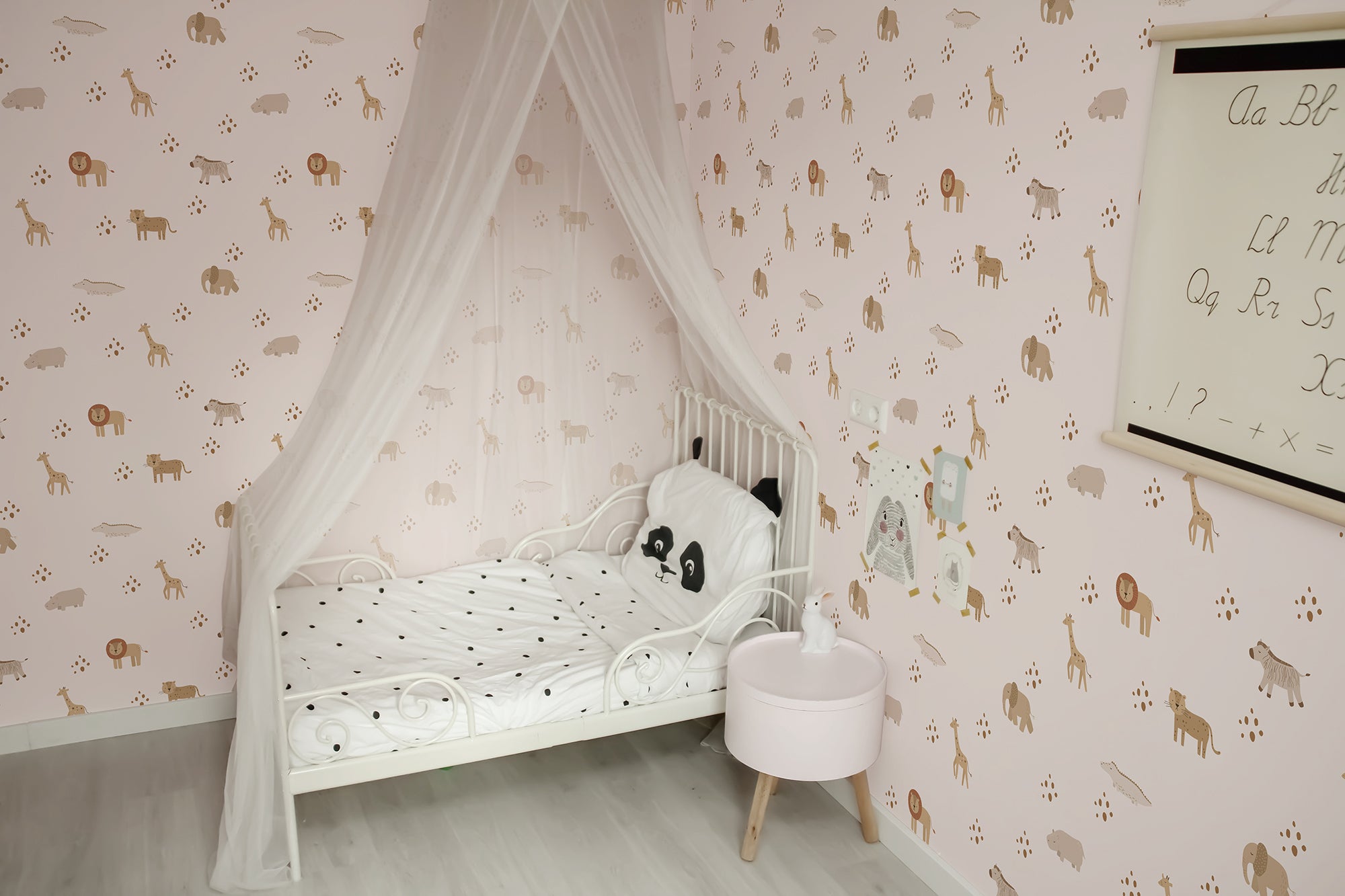 a charming children's bedroom adorned with Safari Animal Wallpaper. This sweet and playful pattern features an array of safari animals such as giraffes, lions, elephants, and zebras in soft brown tones on a pink background. The room is furnished with a white canopy bed with a panda-themed bedding set, and the wall is decorated with whimsical shelves holding toys and educational materials, creating a delightful and imaginative space for a child.