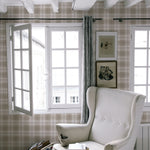 A cozy reading nook illuminated by natural light from large windows, with an elegant wingback chair set against the Winter Plaid Wallpaper. The room's plaid-patterned walls give a feeling of comfort and nostalgia, enhanced by the room's rustic wooden floors and classic decor.