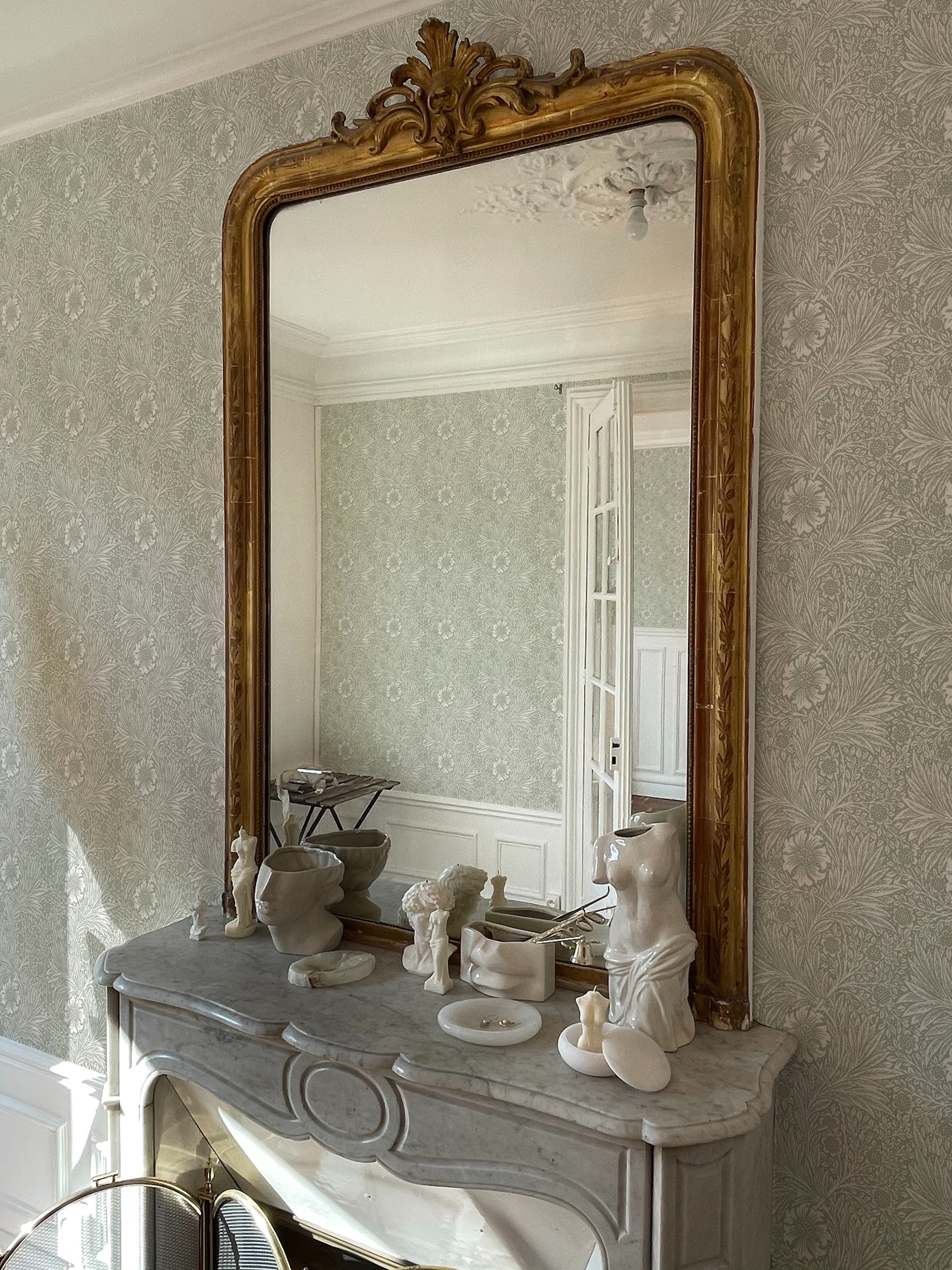 Elegant home office setting showcasing Olive Timeless Floral Wallpaper, featuring a vintage floral pattern in muted olive and beige tones, complementing a classic gold-framed mirror and marble fireplace adorned with porcelain figurines