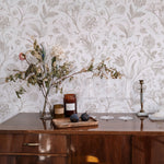 An elegantly appointed room featuring Toile De Jouy Floral Wallpaper, with a detailed print of various flowers and leaves in shades of beige, set against a white backdrop. The wallpaper adds a vintage charm above a wooden sideboard adorned with dried flowers, a candle, and a bottle of wine.